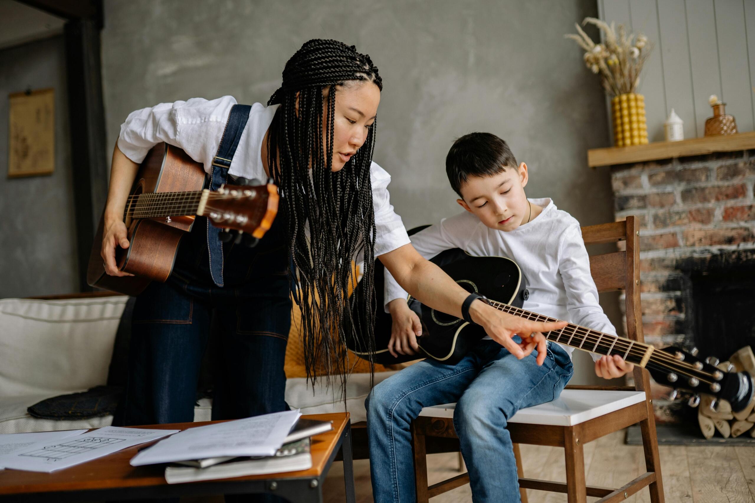 A woman teaching a young boy how to play the guitar, guiding his hands on the strings as they sit together during the lesson