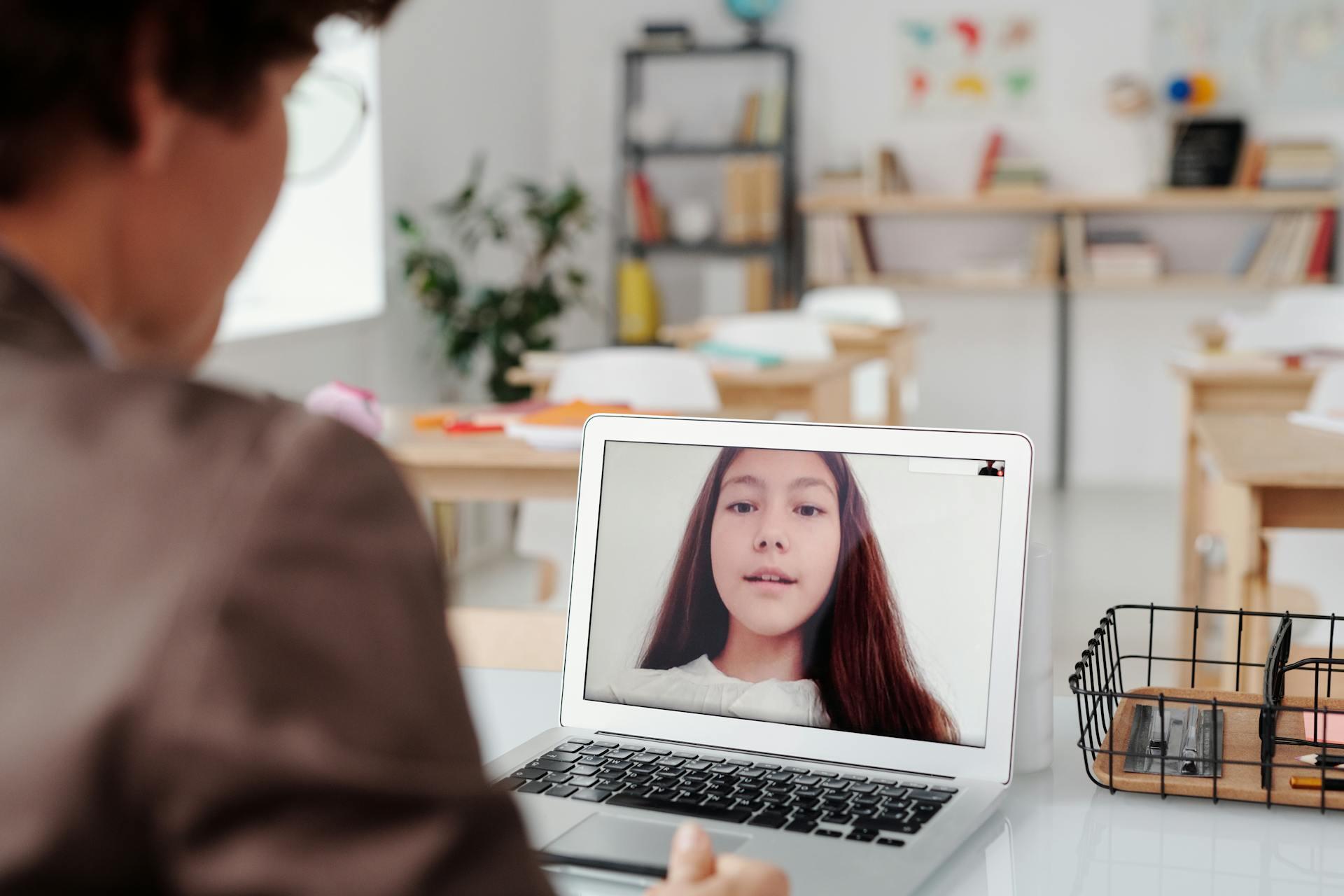 Person video chatting with another person on a laptop in a bright, organized room with desks and shelves in the background.