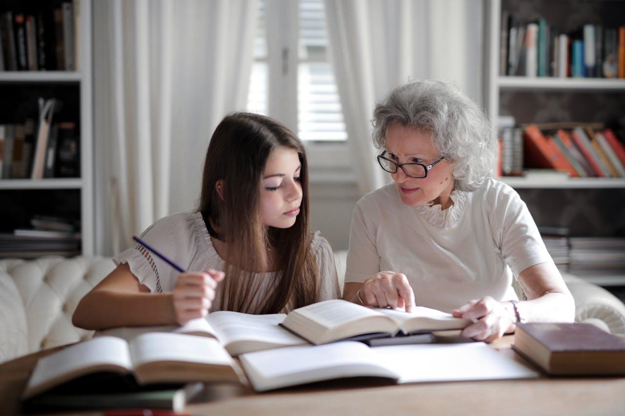 A young girl and an older woman are engaged in reading and studying together at a table filled with books in a cozy room.