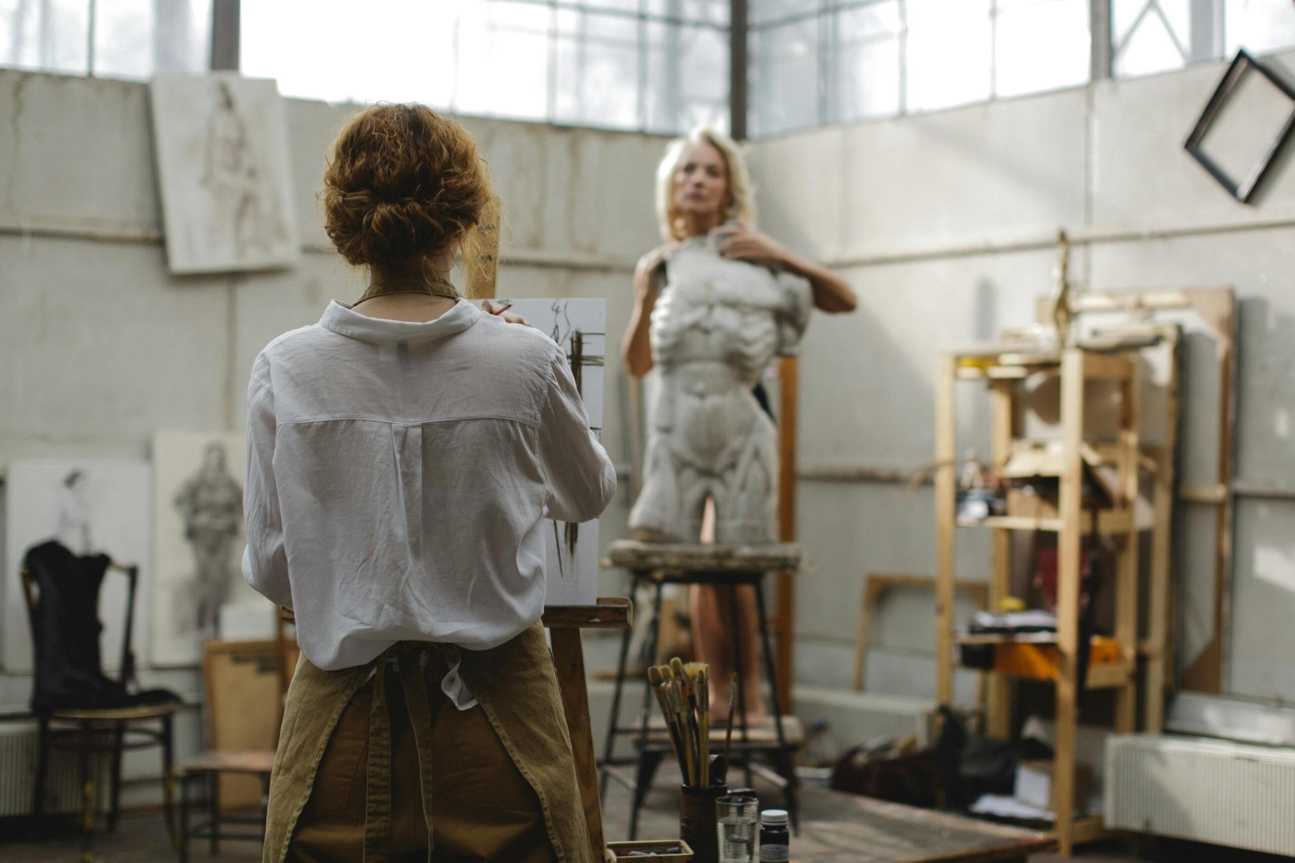 A life drawing model posing in a well-lit studio while an artist sketches from an easel nearby