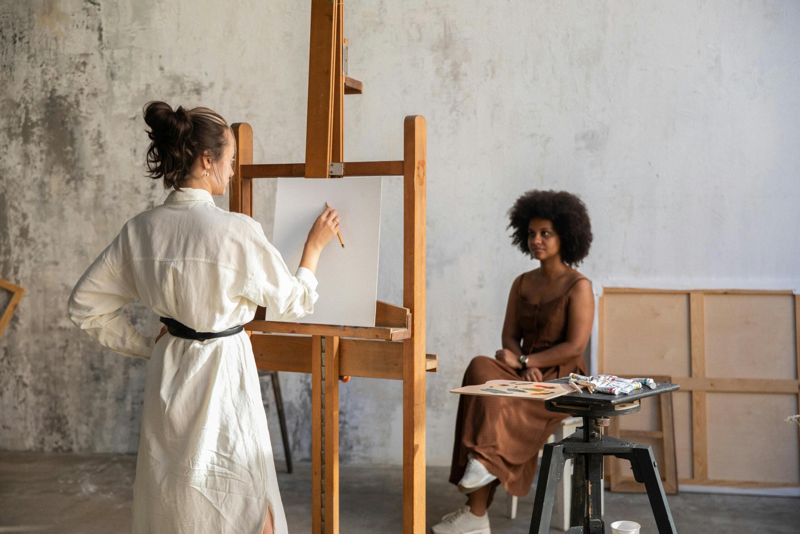 A woman seated at an easel sketching while a life model poses in a dress, sitting calmly in a chair across from her.
