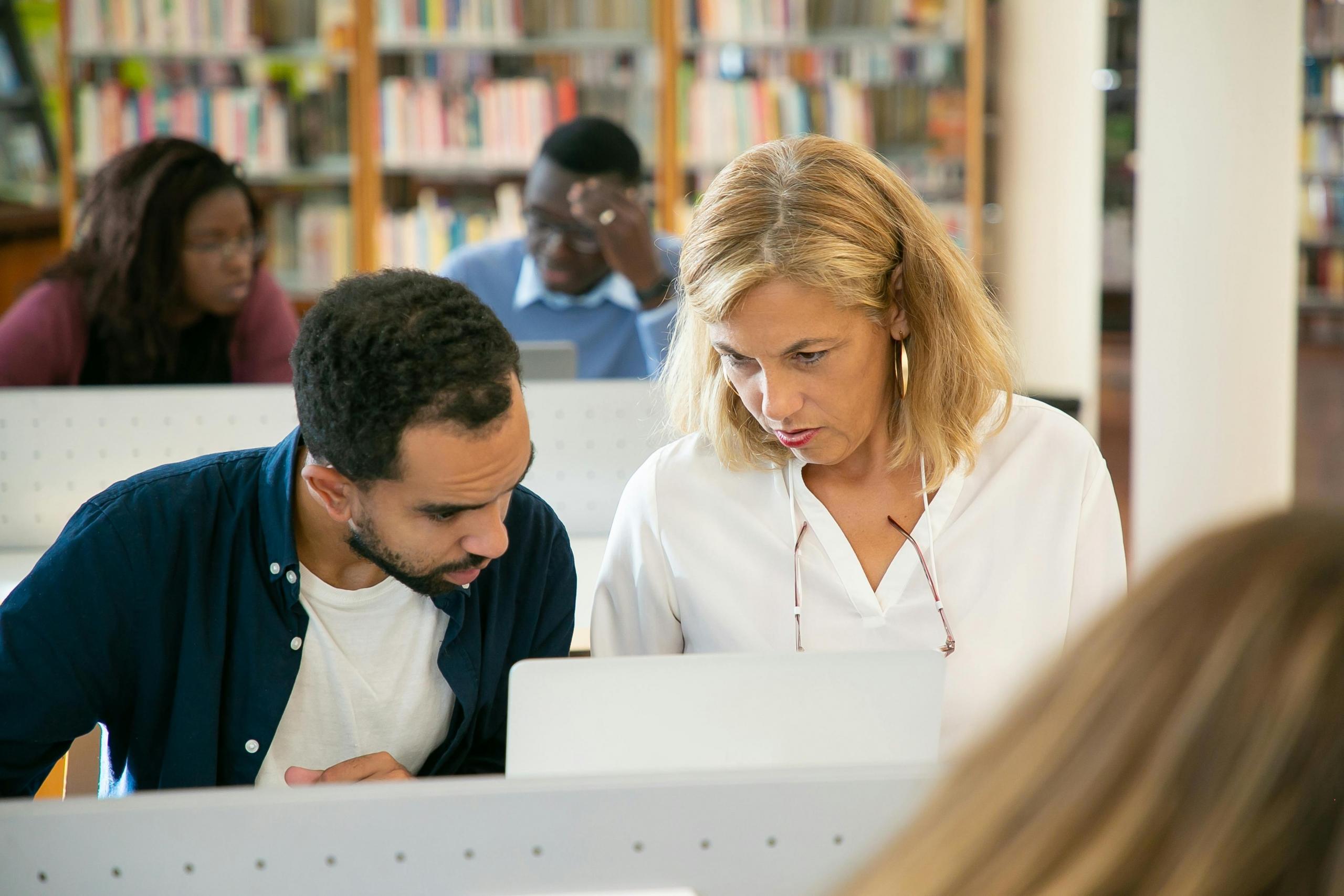 Two individuals sit closely at a desk in a library, collaborating over a laptop, surrounded by shelves filled with books.