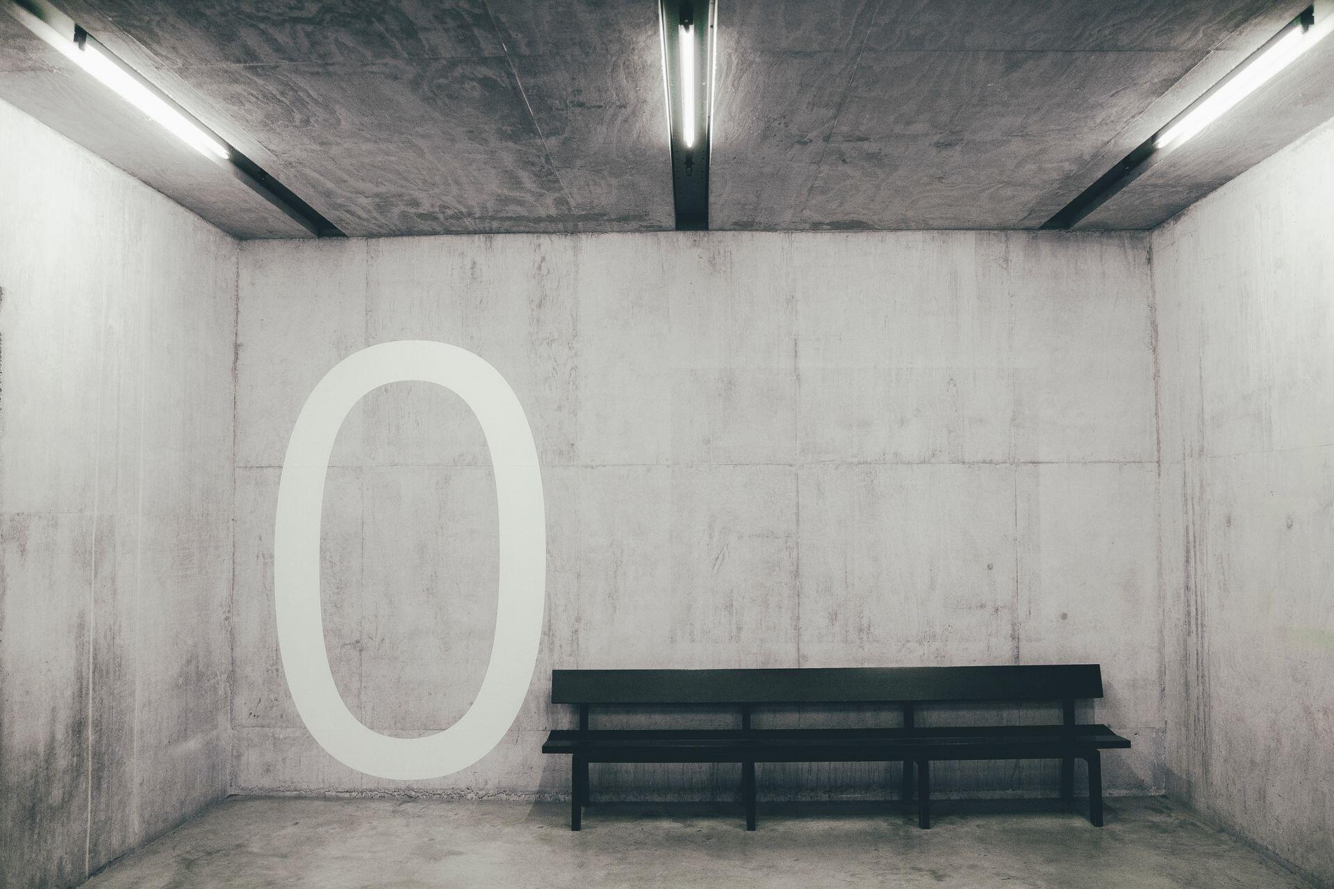 A black bench sits in a bare concrete room beside a large white numeral zero painted on the wall.