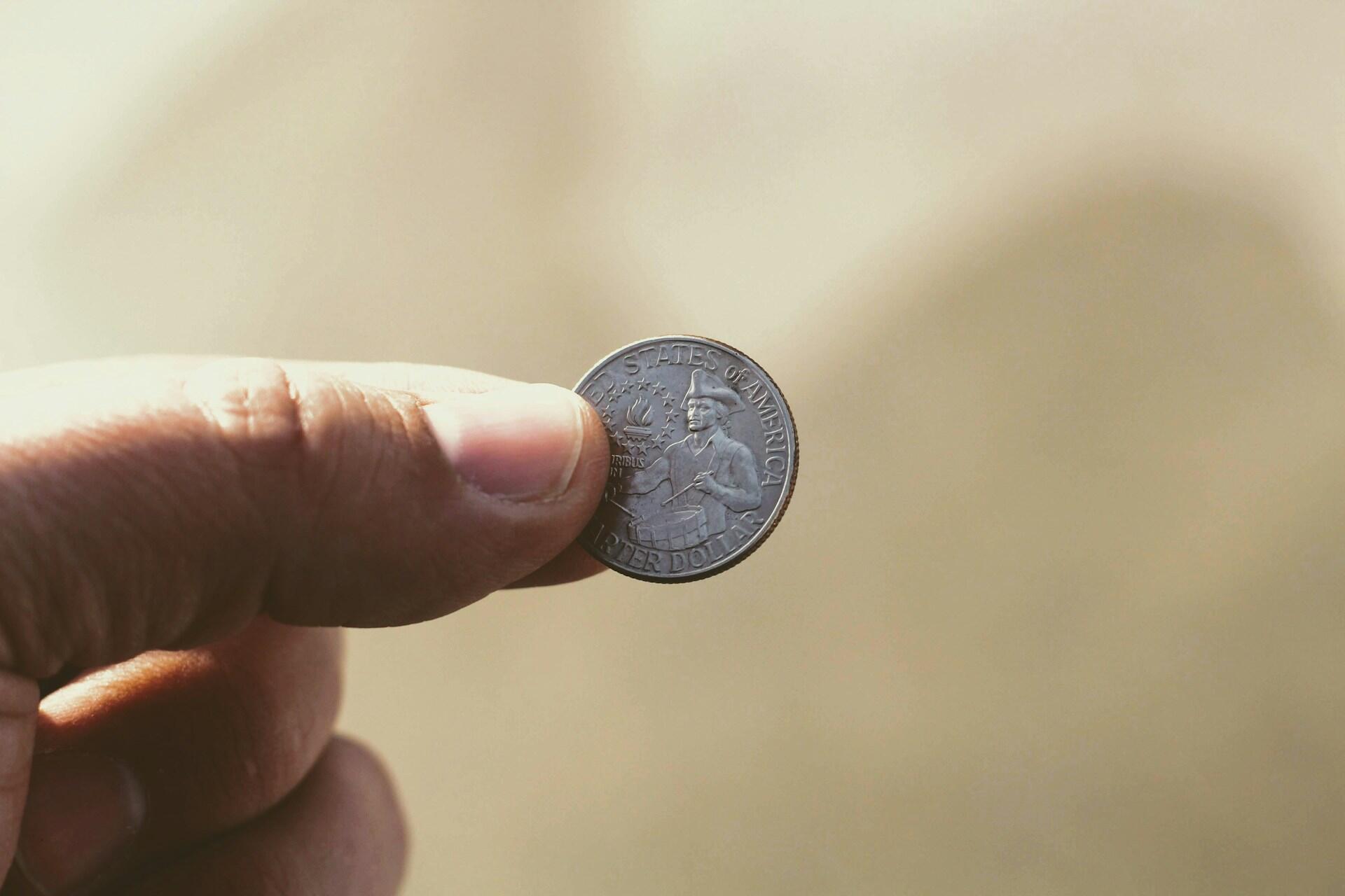 Close-up of a thumb and forefinger holding a United States quarter dollar coin featuring a colonial figure against a soft, warm beige background.