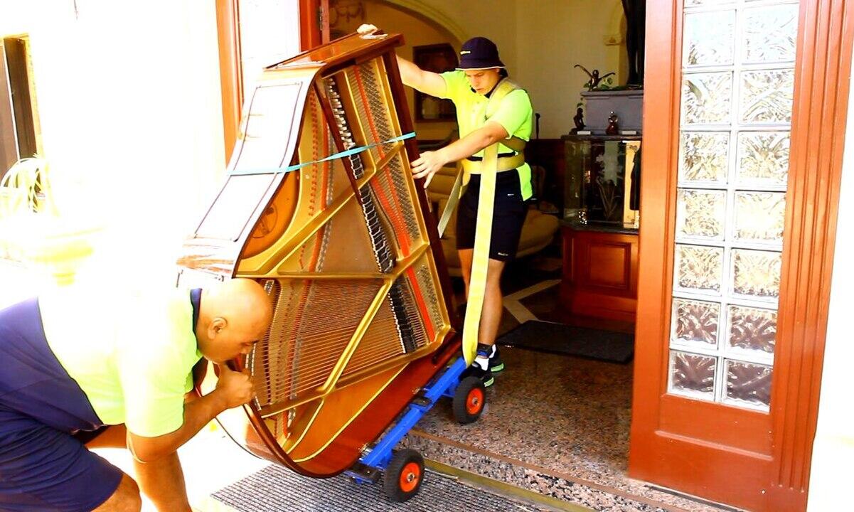 Two movers in bright shirts carefully transport a grand piano out of a house, using a dolly and securing it with straps.