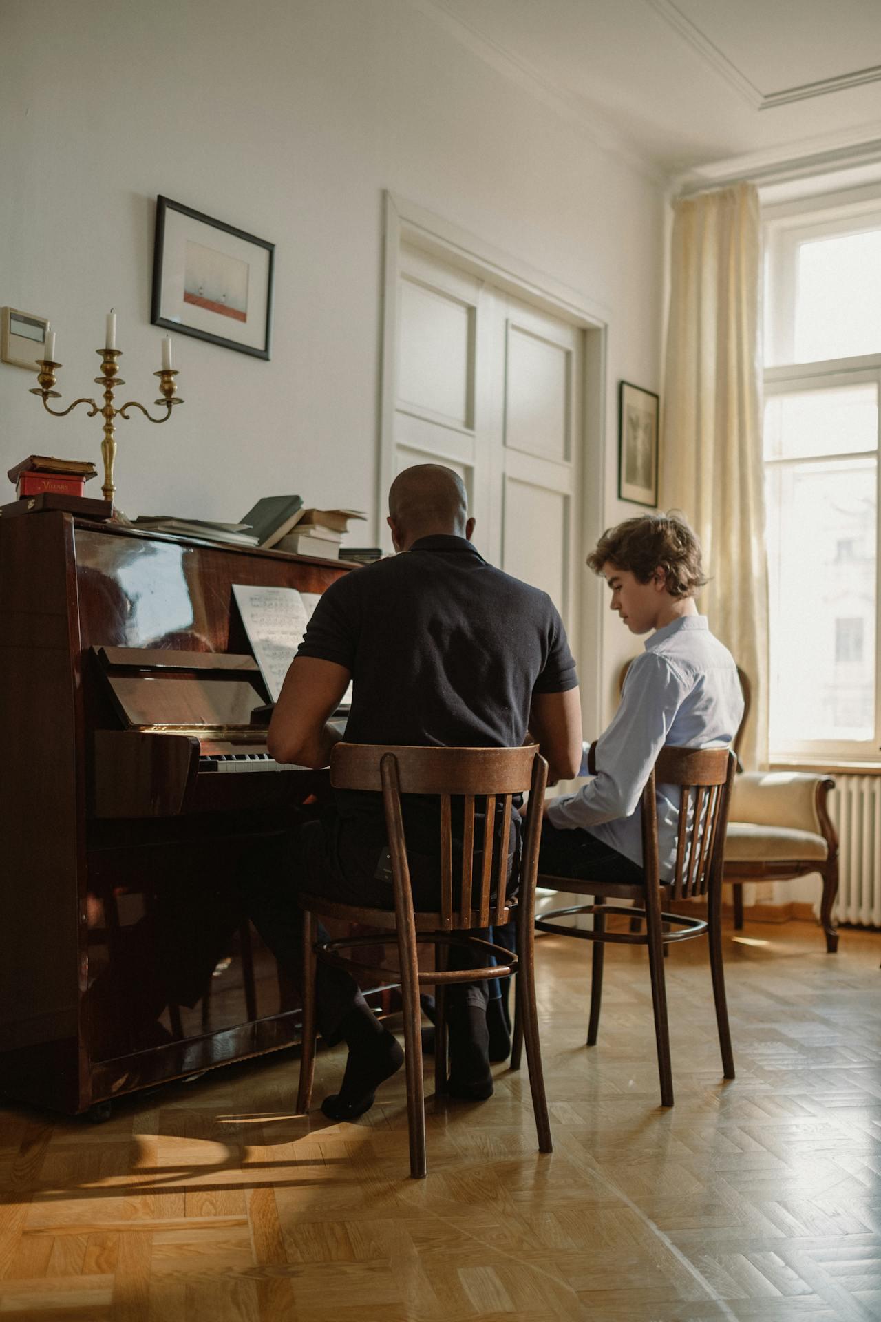 A person teaches piano, sitting at an upright piano, while another student attentively listens, in a sunlit, cozy room.
