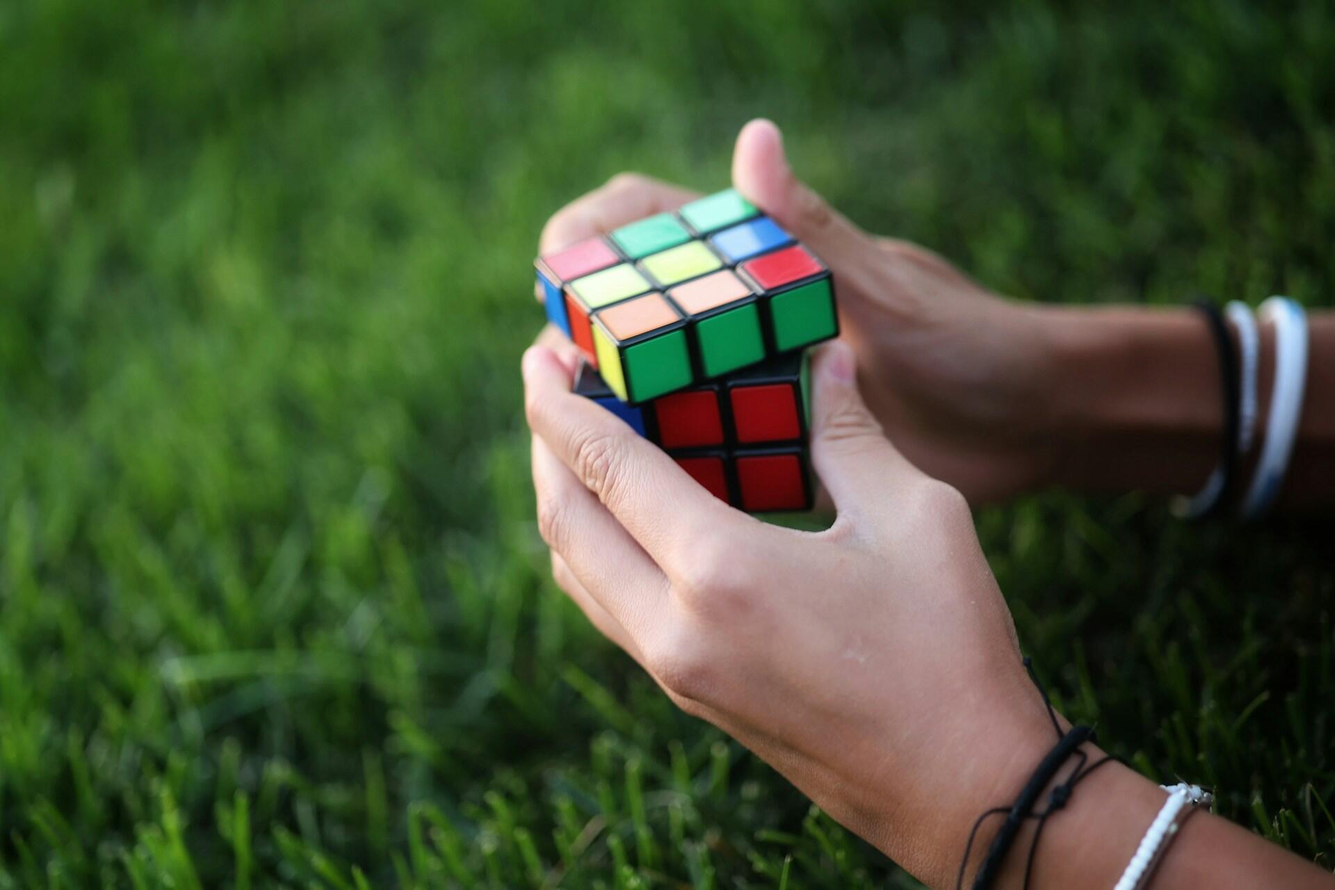 Two hands holding and twisting a colorful, partially solved Rubik's Cube against a blurred background of green grass.