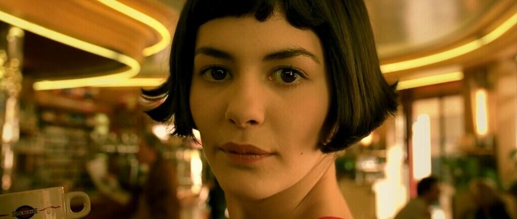 A person with short dark hair in a warmly lit cafe with curved ceiling lights and blurred patrons in the background.