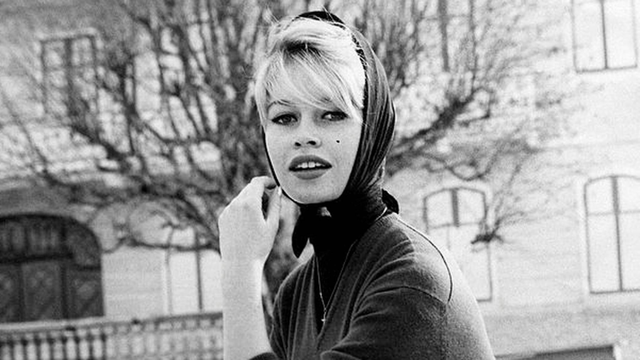 Black and white photo of a person wearing a headscarf and cross necklace, standing outdoors with bare trees and a building in the background.