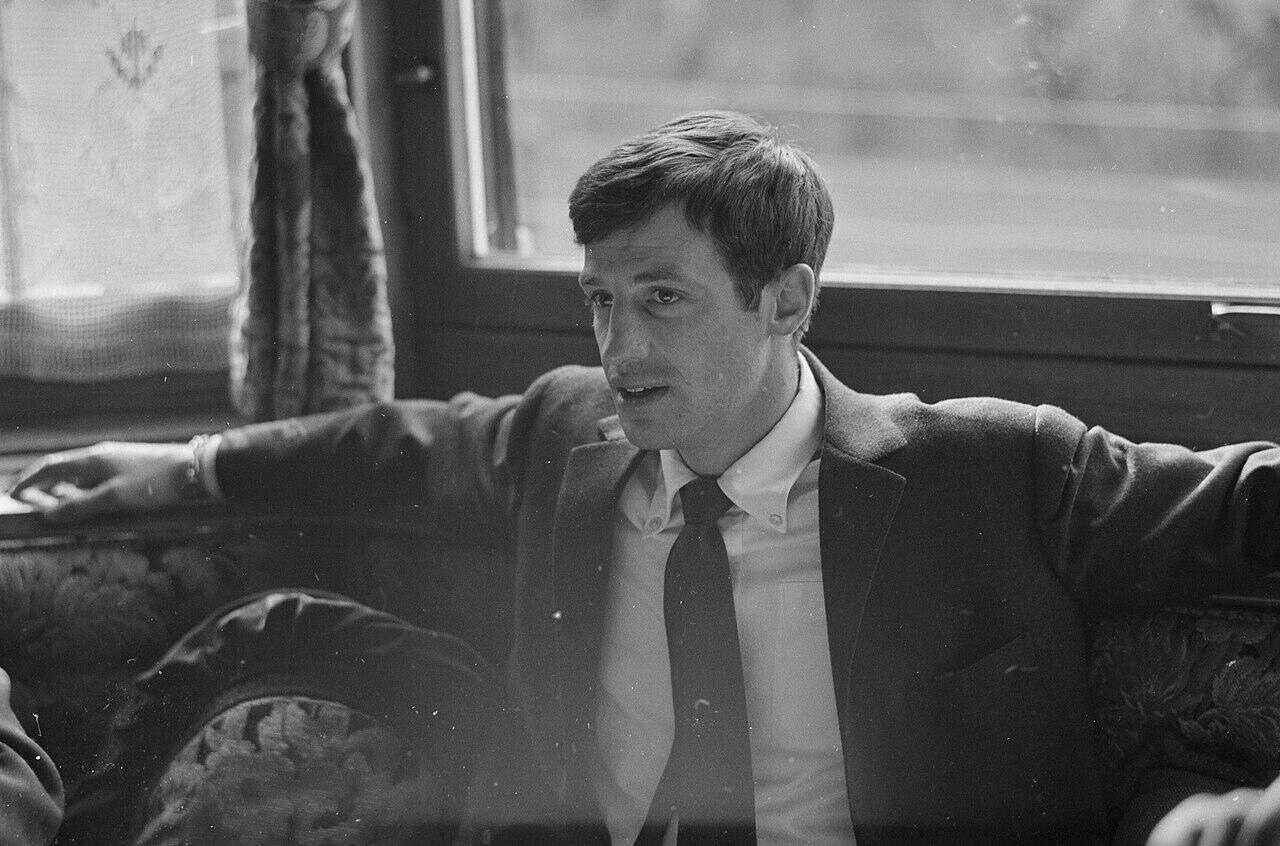 Black-and-white photo of a man in a suit and tie sitting with arms spread on a cushioned bench near a window with curtains.