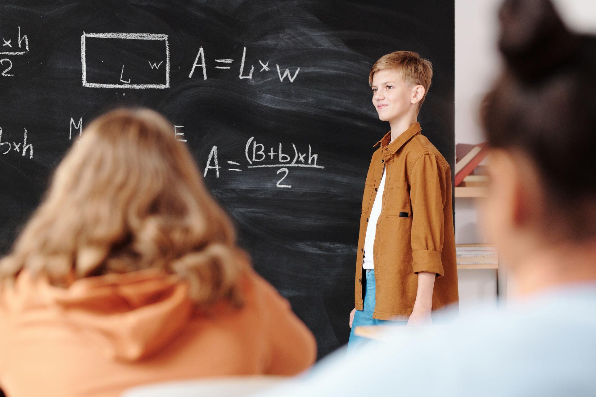 A boy stands by a blackboard with math formulas as two classmates are partially visible in the foreground.