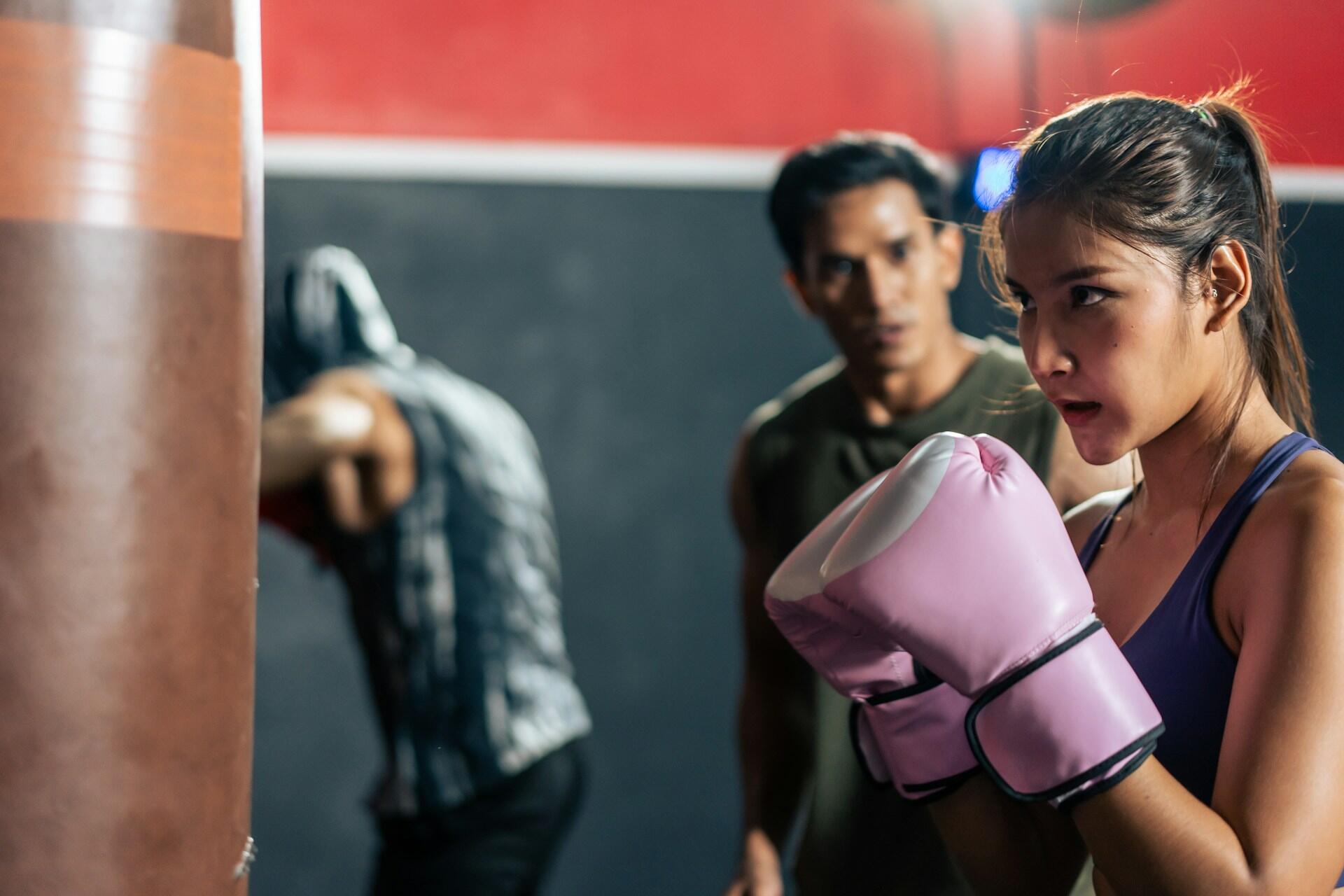 Woman wearing pink boxing gloves training in a gym with a man and another person punching a bag in the background.