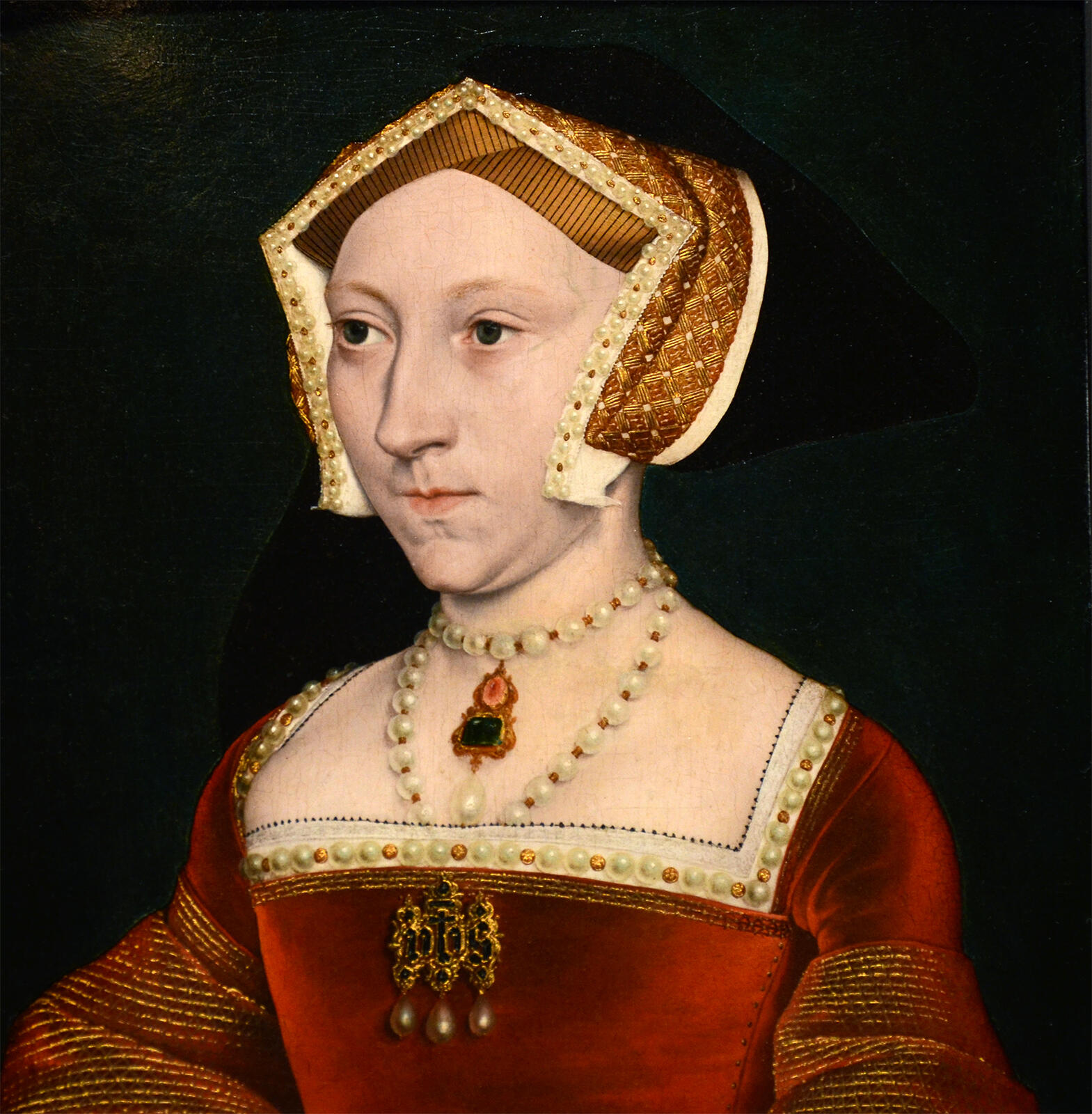 A woman in a richly detailed red dress adorned with pearls and gold jewelry, wearing an elaborate headdress against a dark background.