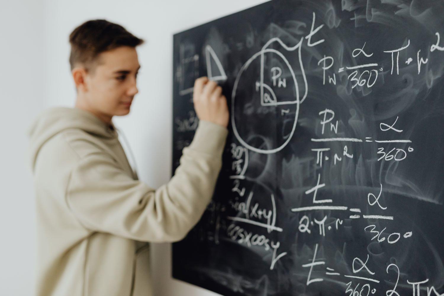 A person in a hoodie writes mathematical formulas on a black chalkboard covered with various equations and diagrams.