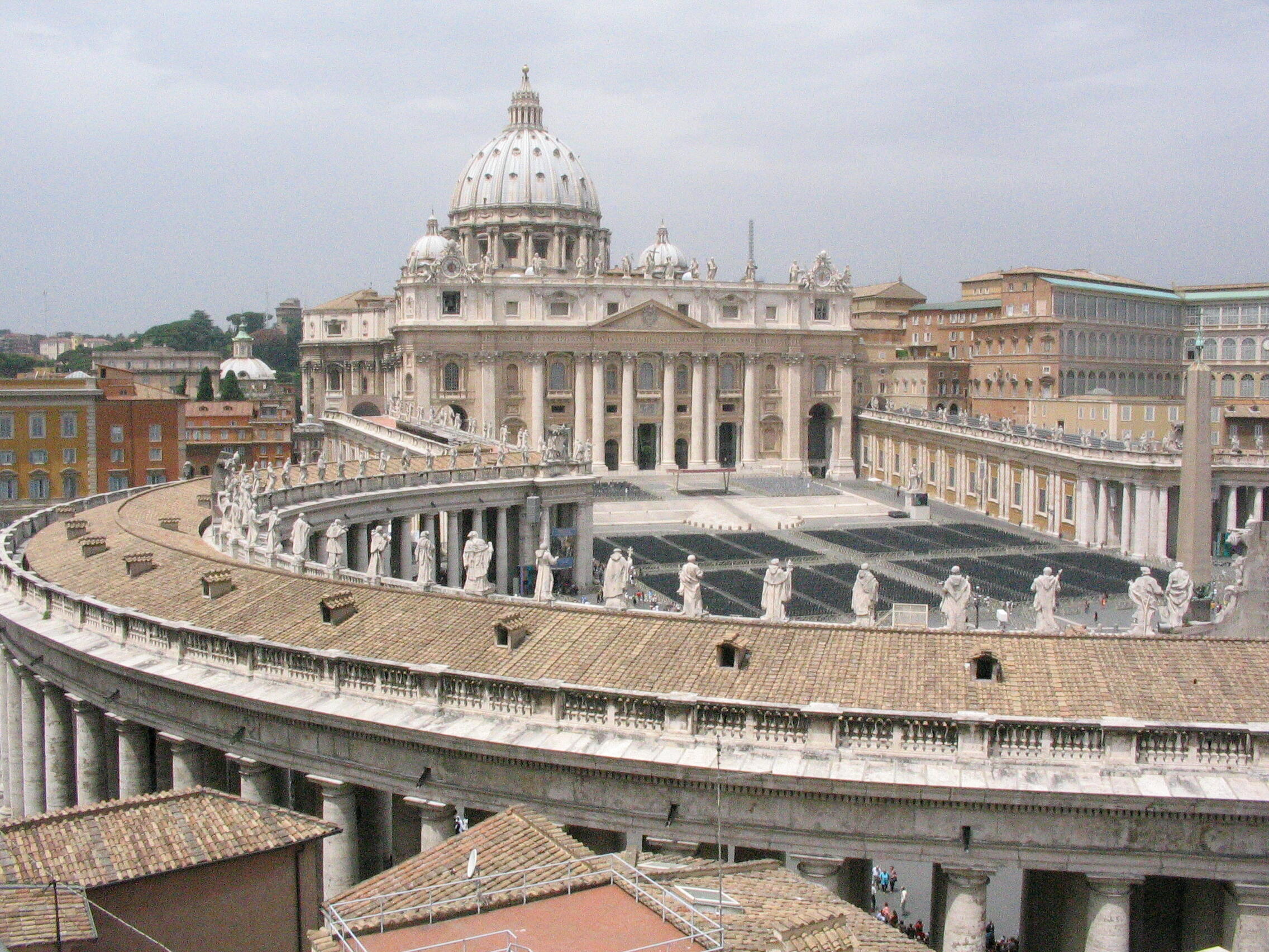 St. Peter's Basilica in Vatican City, showcasing its grand dome, colonnades, and statues under a cloudy sky.