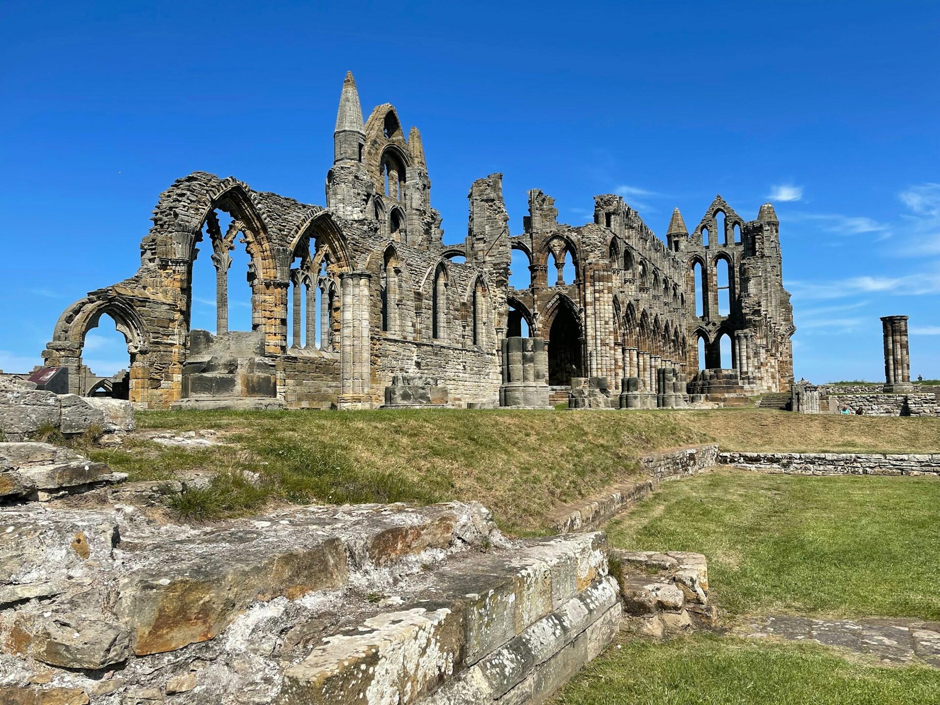 Ruins of an ancient abbey rise against a clear blue sky, showcasing Gothic arches and stone walls amidst green grass.