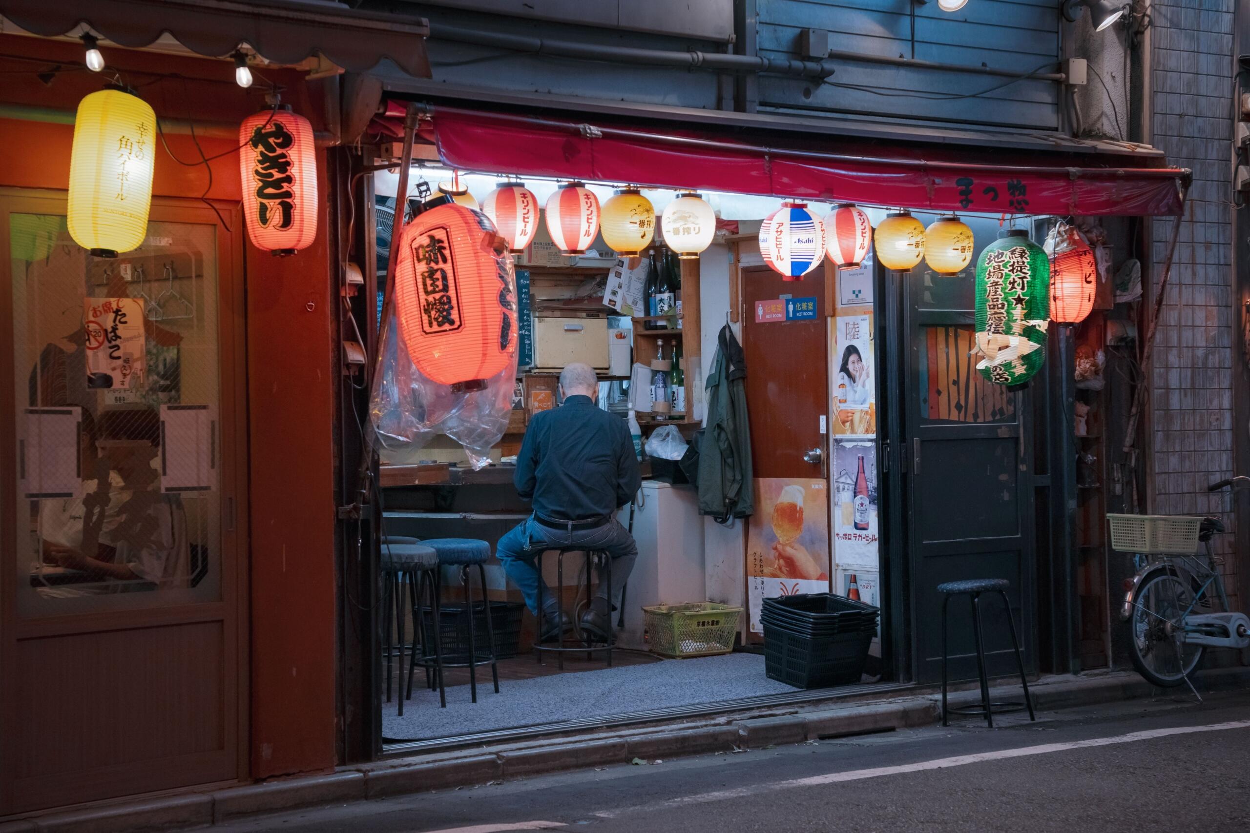 A small Japanese diner in Tokyo with warm lighting, counter seating, and a cozy interior. Wooden details, simple stools, and shelves of dishes create an intimate late-night atmosphere, suggesting a welcoming spot for ramen, rice bowls, and quiet conversation.