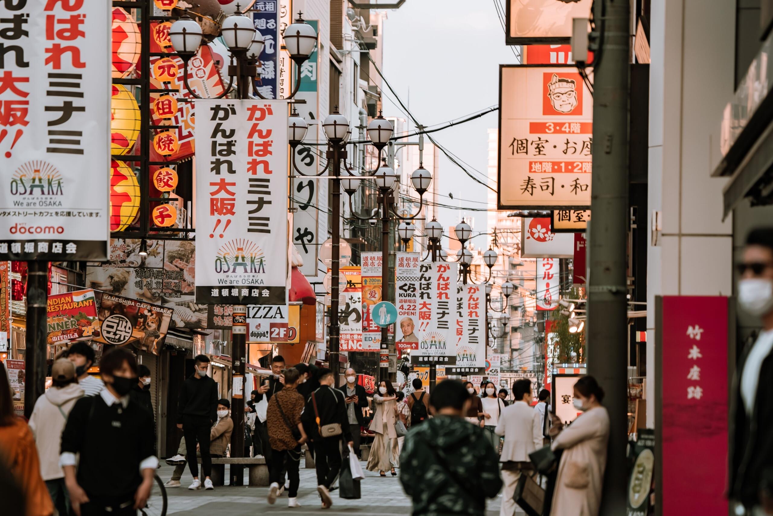 A bustling street in Japan lined with brightly lit storefronts and towering buildings covered in colorful neon signs and illuminated billboards.