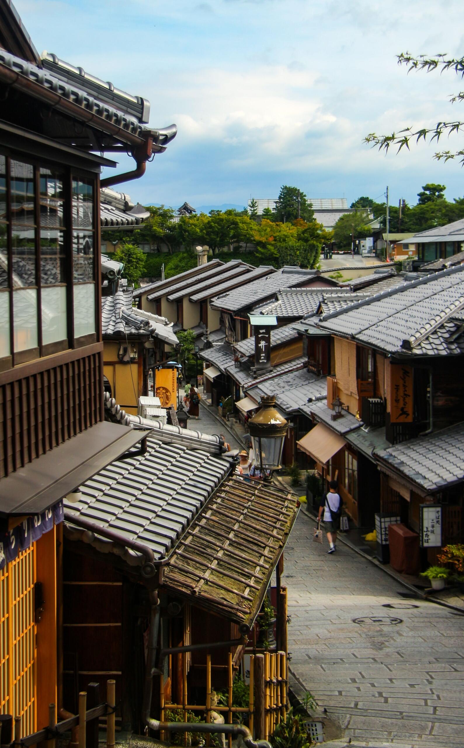 A traditional street in Kyoto lined with historic wooden buildings, stone-paved walkways, and classic Japanese storefronts. Paper lanterns and tiled roofs reflect the city's preserved architecture.