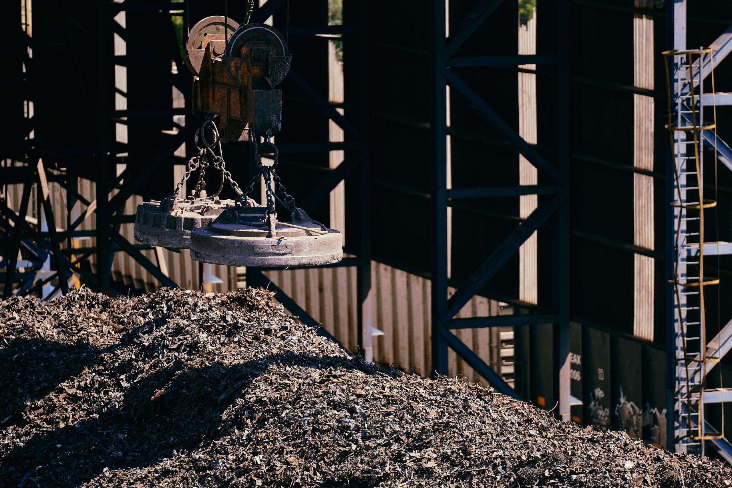 A large industrial electromagnet lifts scrap metal in a junkyard, with piles of discarded metal and machinery in the background.