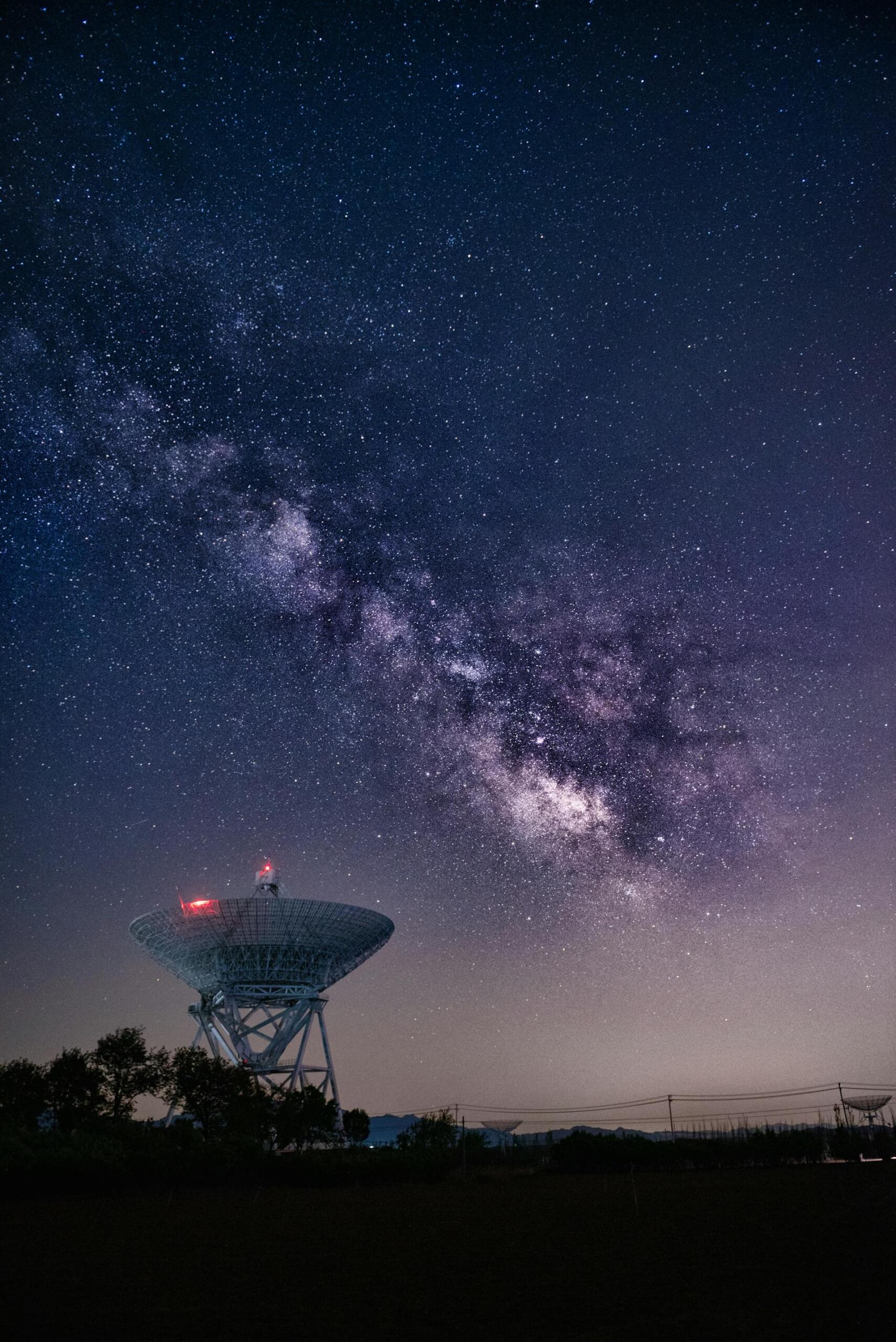 A large satellite dish stands under a starry sky, with the vibrant Milky Way galaxy glowing above in a stunning cosmic display.