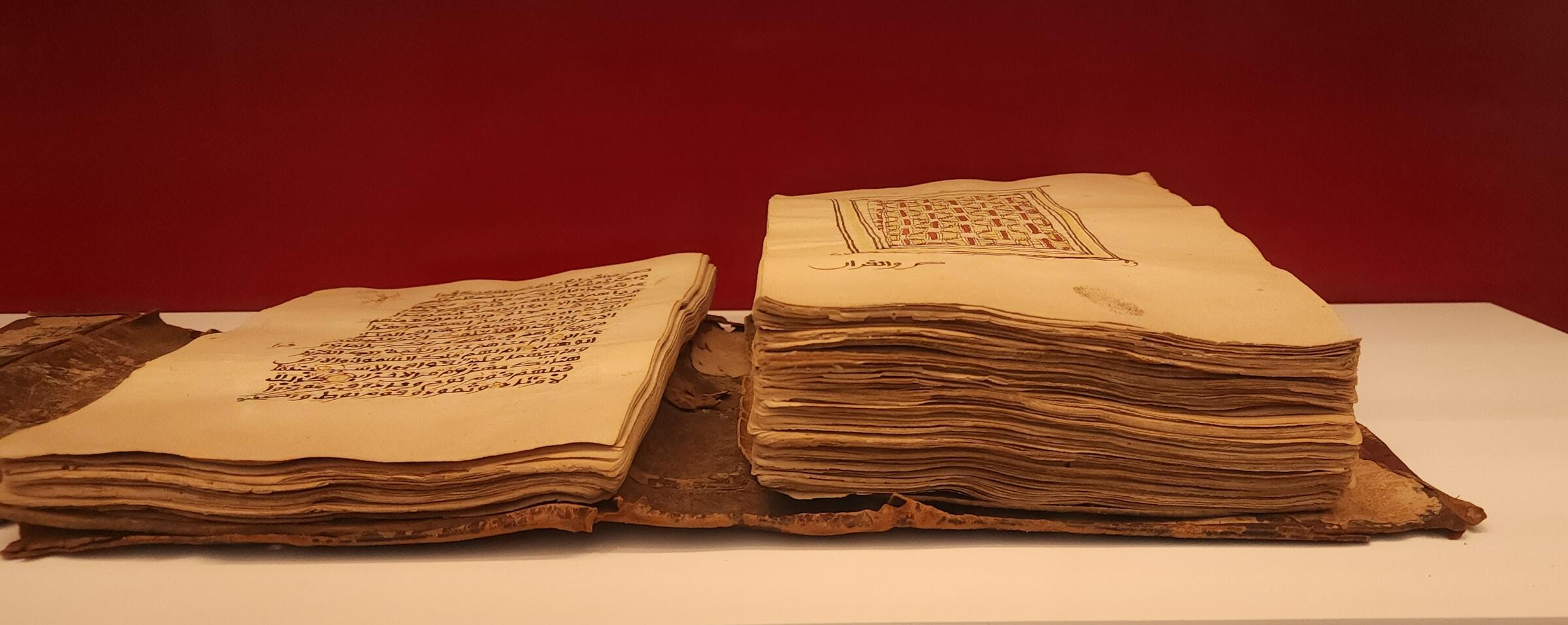 Old, thick manuscript with Arabic script and ornate design on worn pages, displayed on a white surface against a red background.