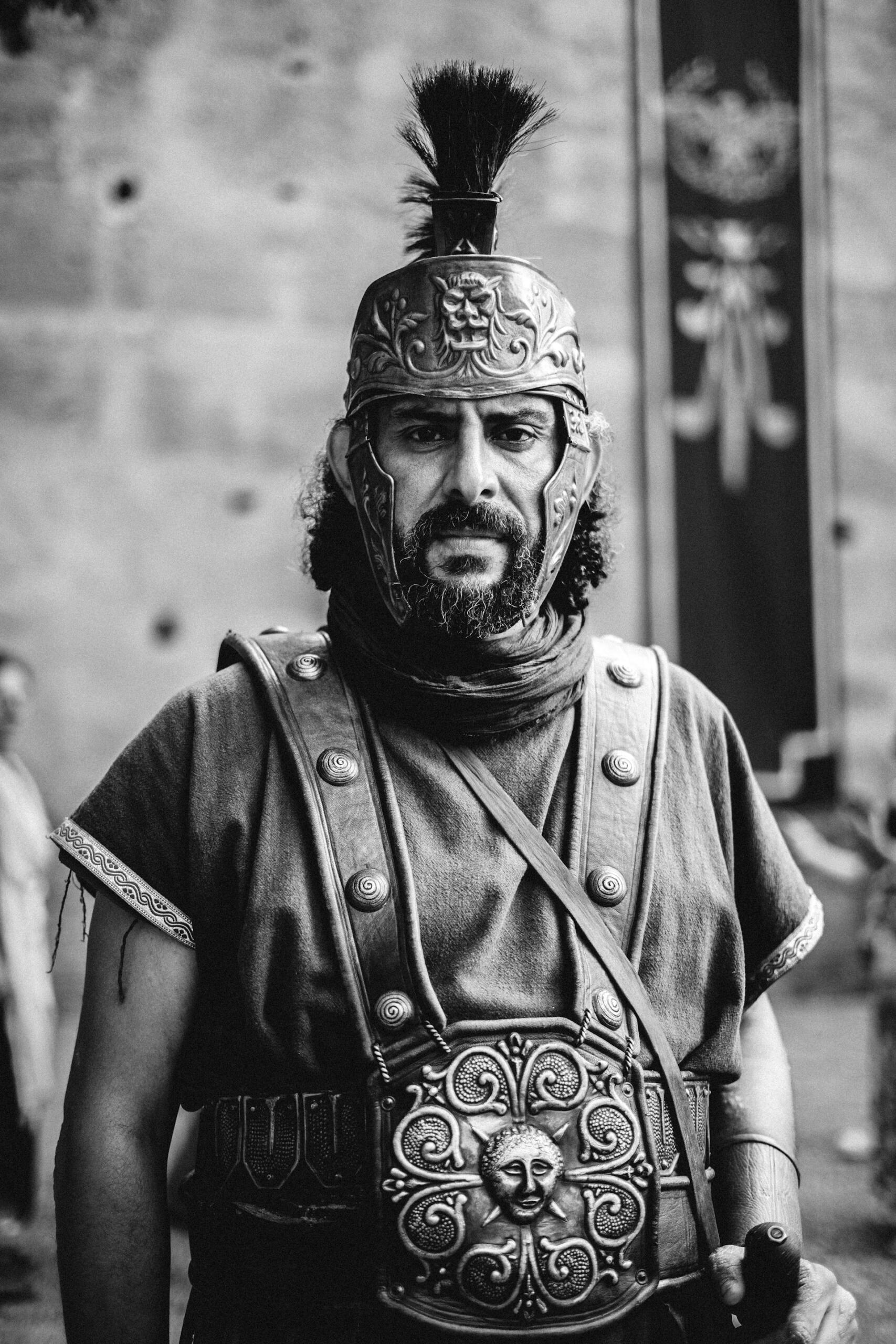 Black and white photo of a person dressed as a Roman soldier, wearing detailed armor and helmet with plume, standing indoors.