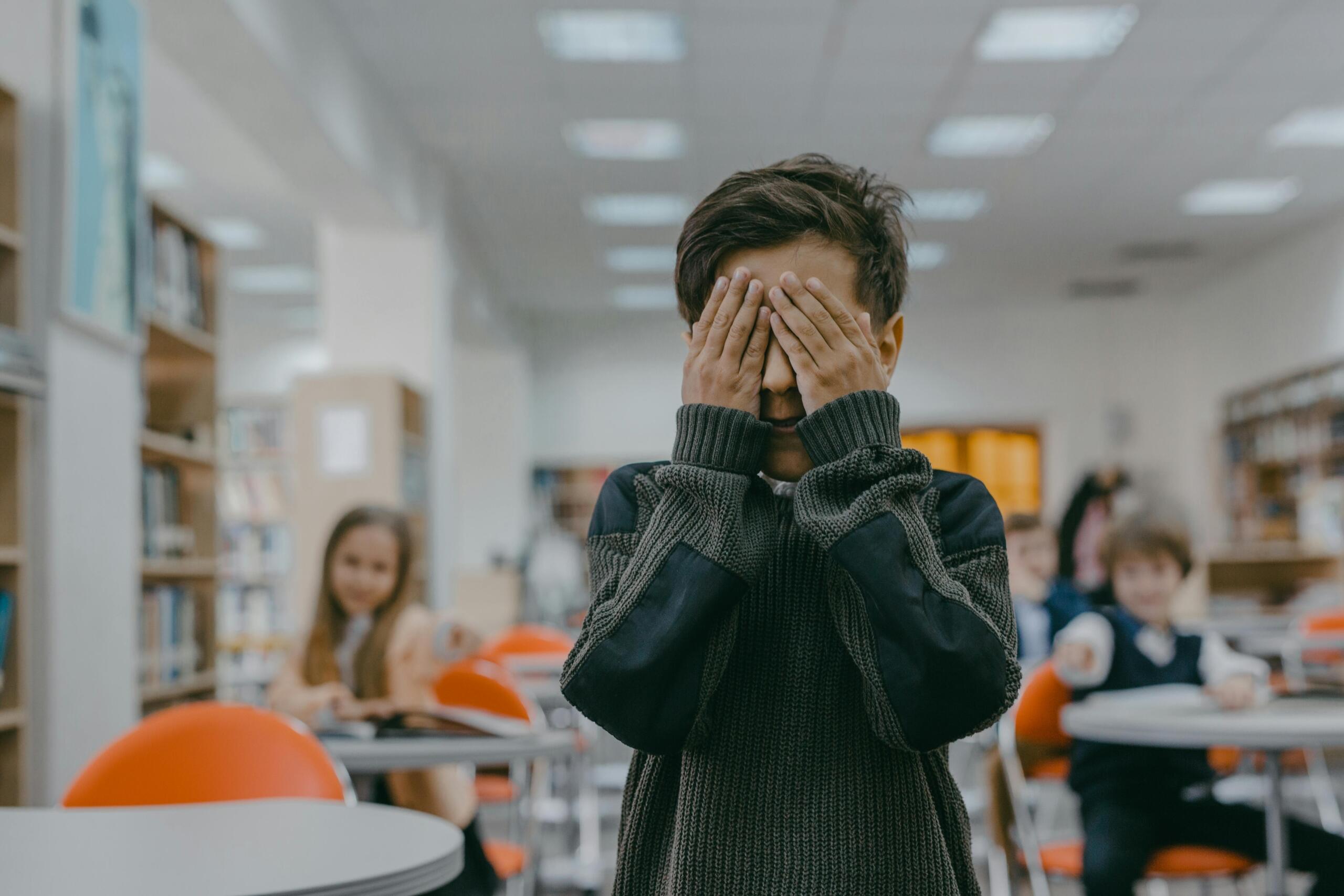 A child covers his face in a classroom while children point at laugh at him.