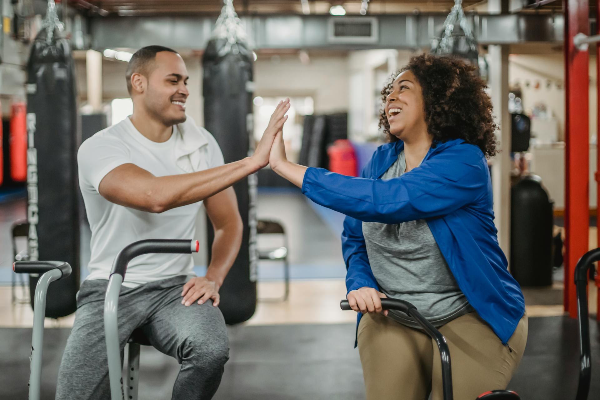 Two people face each other in a gym, engaging in a fun high-five gesture while sitting on exercise equipment.