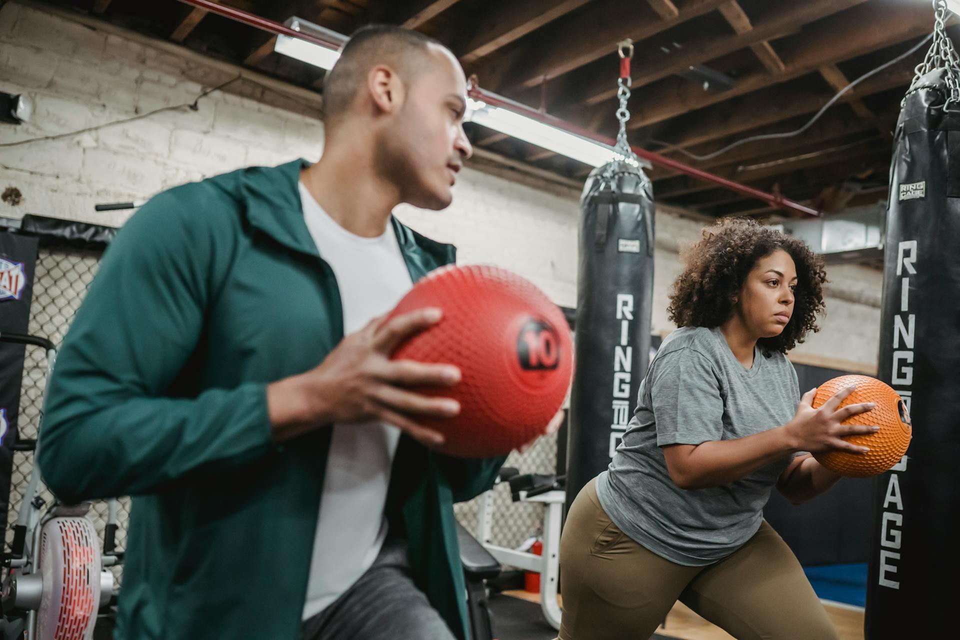 Two individuals engaged in a workout, each holding a medicine ball, in a gym with boxing bags and fitness equipment in the background.