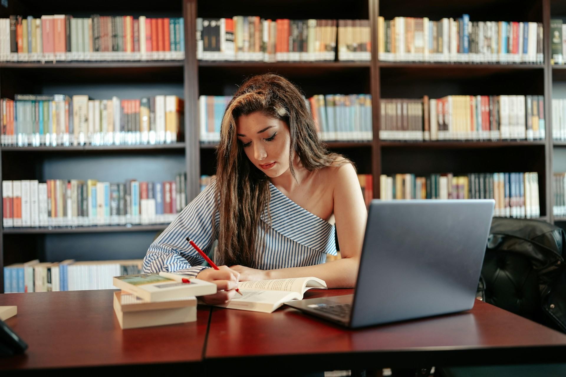 Person studying with books and laptop at a library desk with a bookshelf in the background.