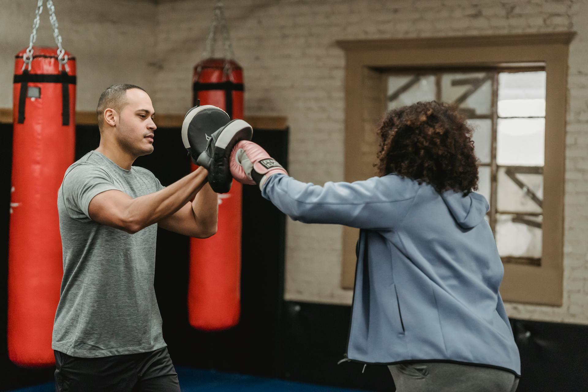 A person in boxing gloves practices punches with a trainer holding focus pads in a gym adorned with red punching bags.