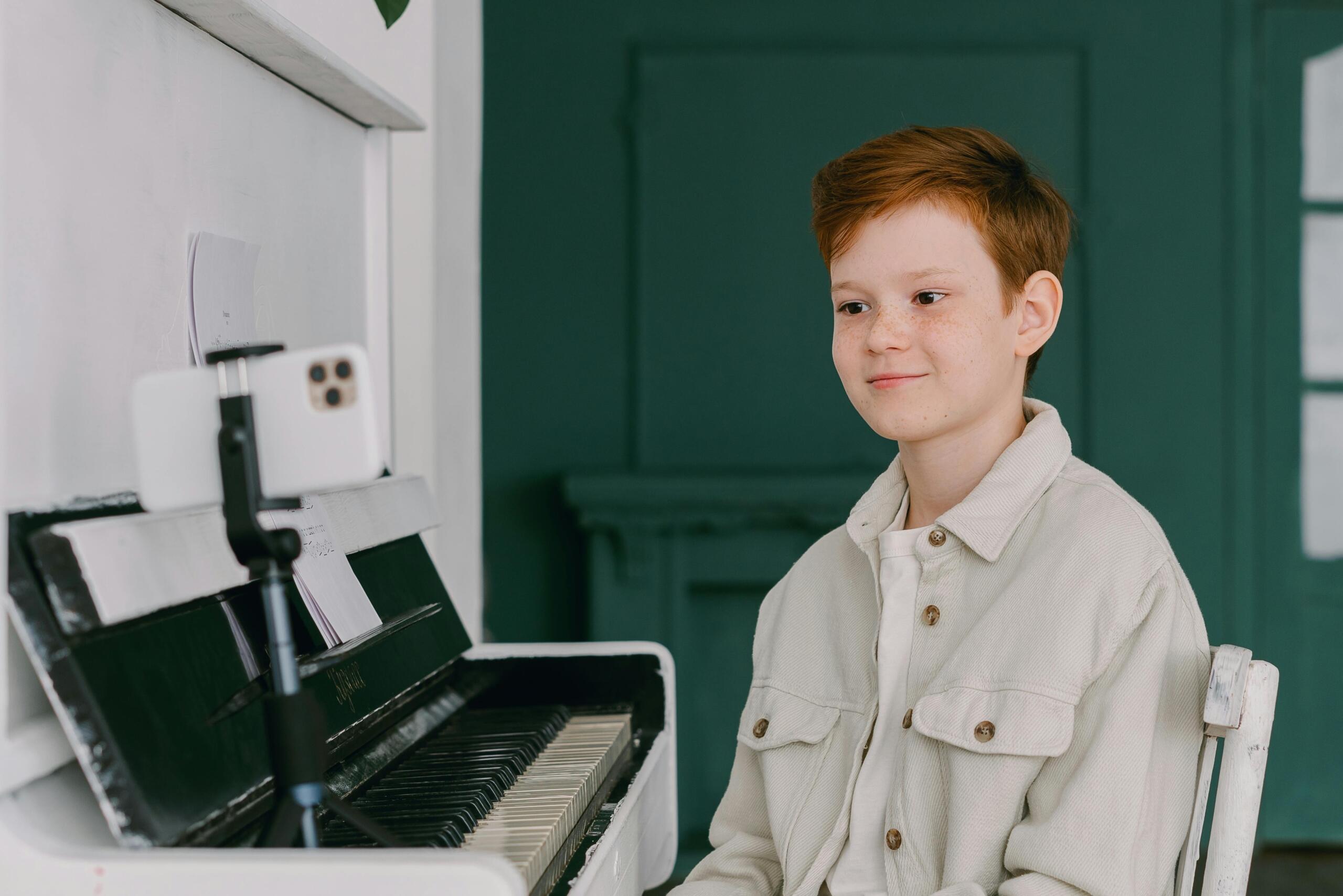 A boy sitting at an upright piano smiles at a smartphone, using it to learn or record music.