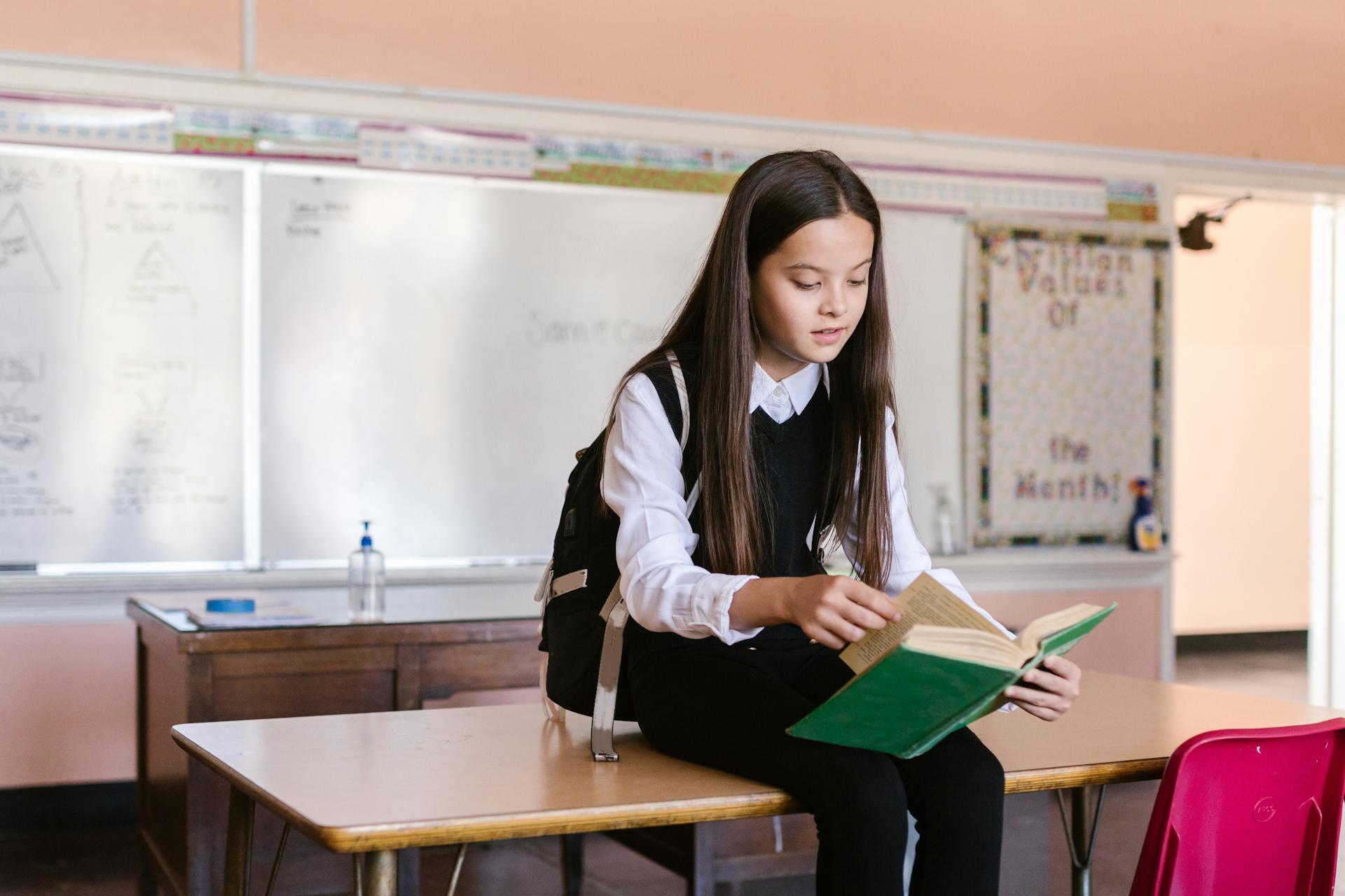 A student in a black vest and white shirt sits on a wooden table, reading a green book, in a classroom setting.