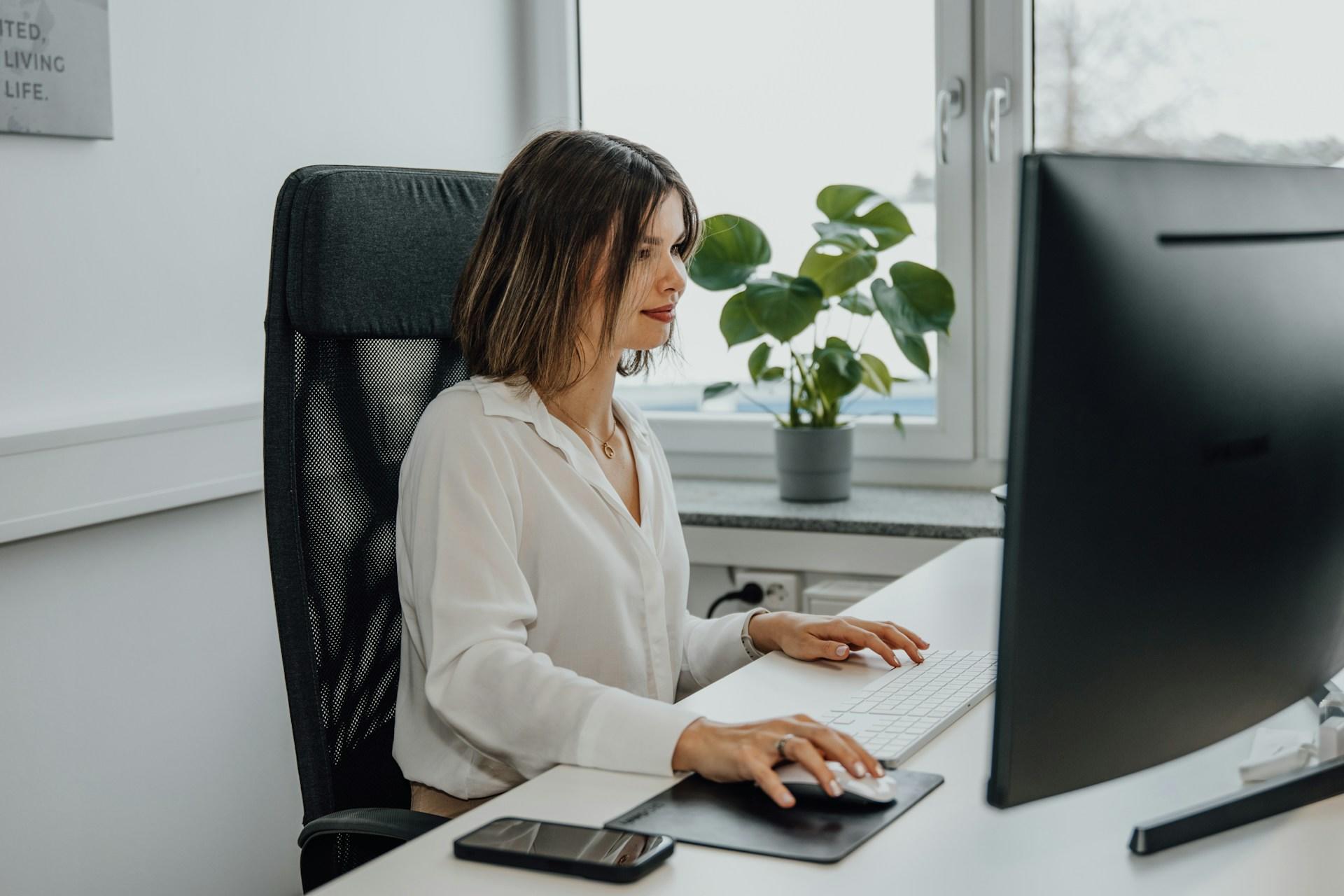 Person at a desk with a computer, keyboard, mouse, and smartphone, near a potted plant.
