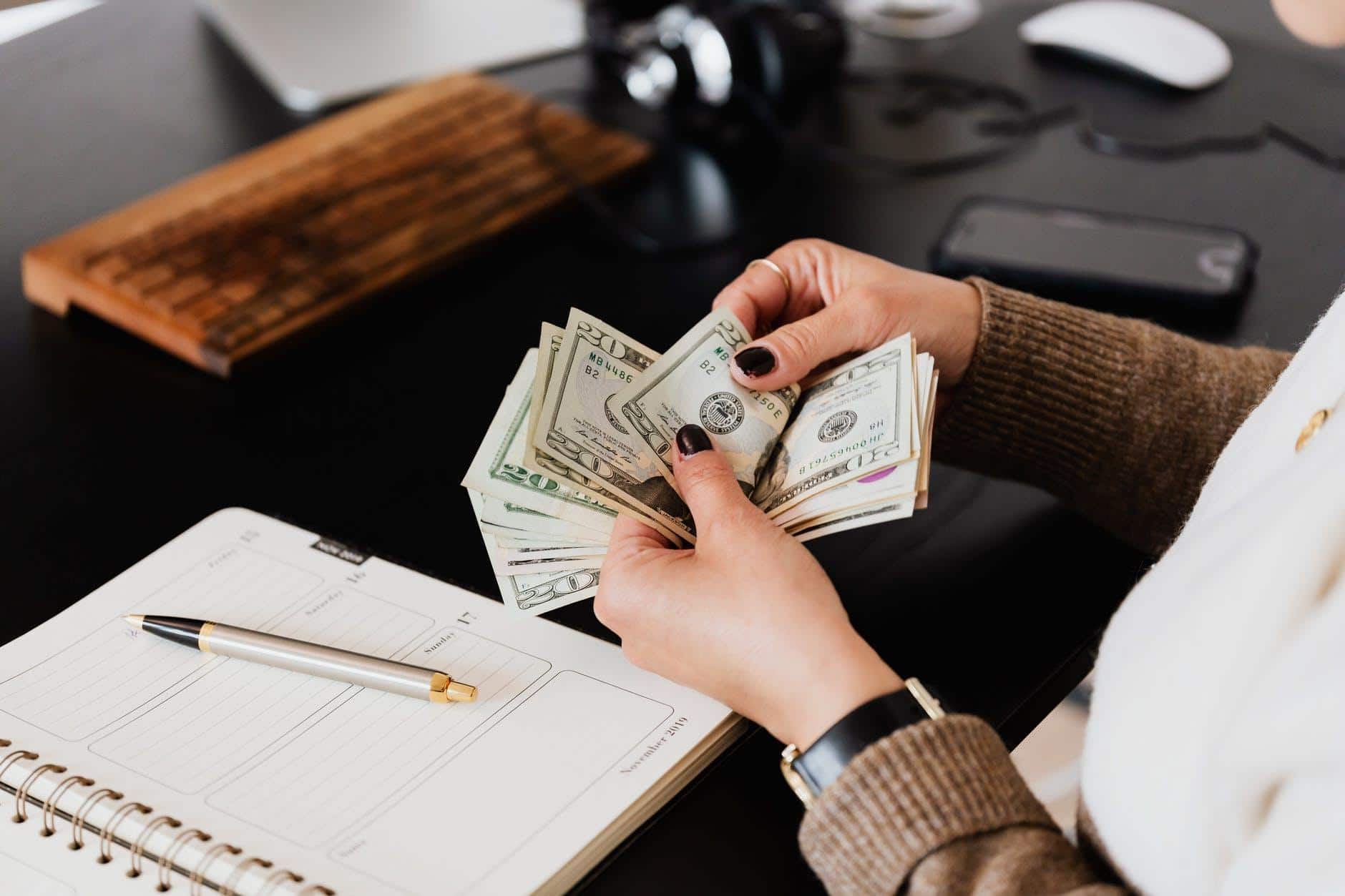 The photograph shows the hands of a person counting money bills on a desk.
