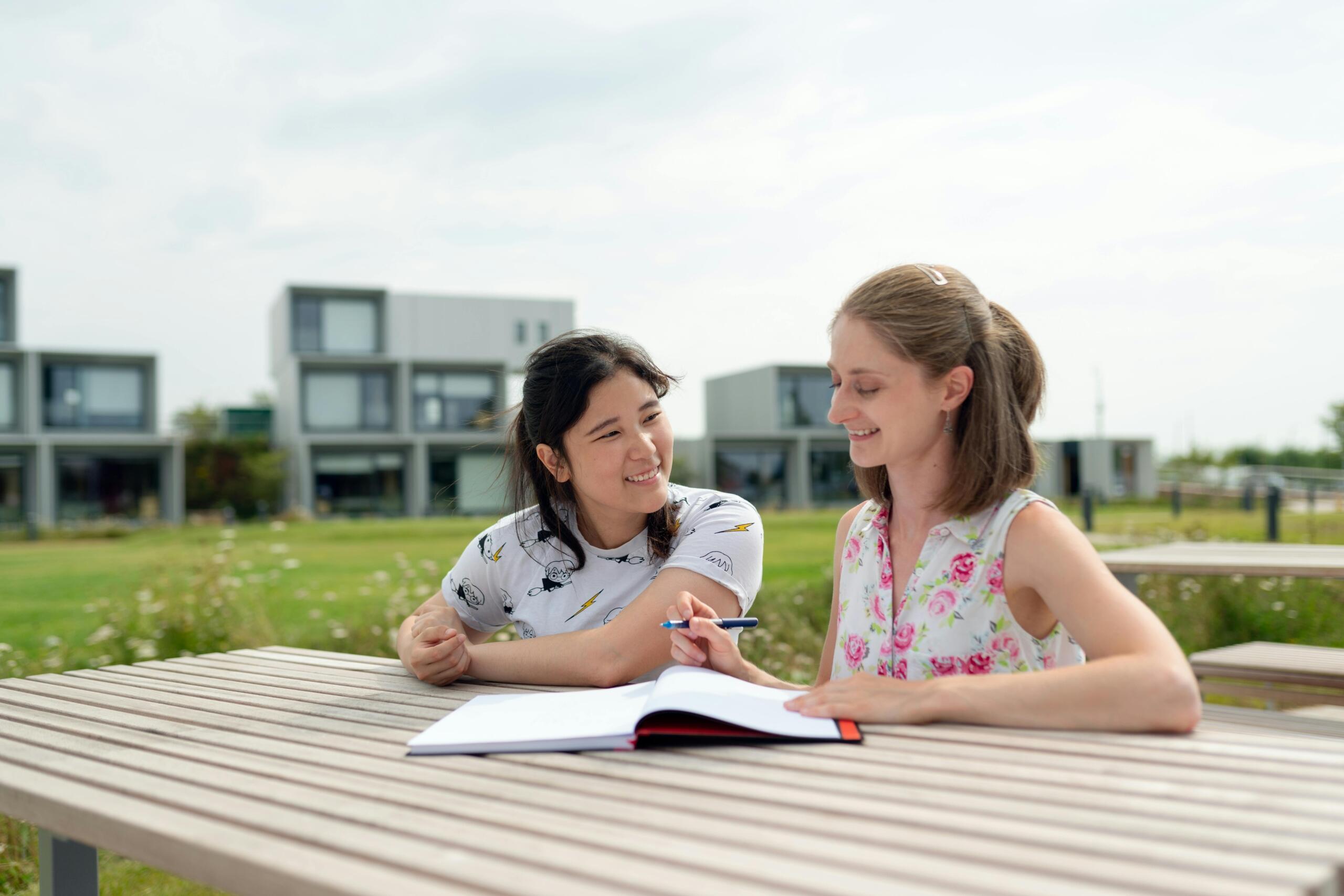 Two young women sitting outside at a wooden table, smiling and studying together with an open notebook, in front of a row of modern buildings on a sunny day.
