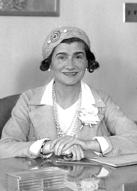 Coco Chanel sits at a table wearing a hat, pearl necklace, and jacket, with her hands clasped.