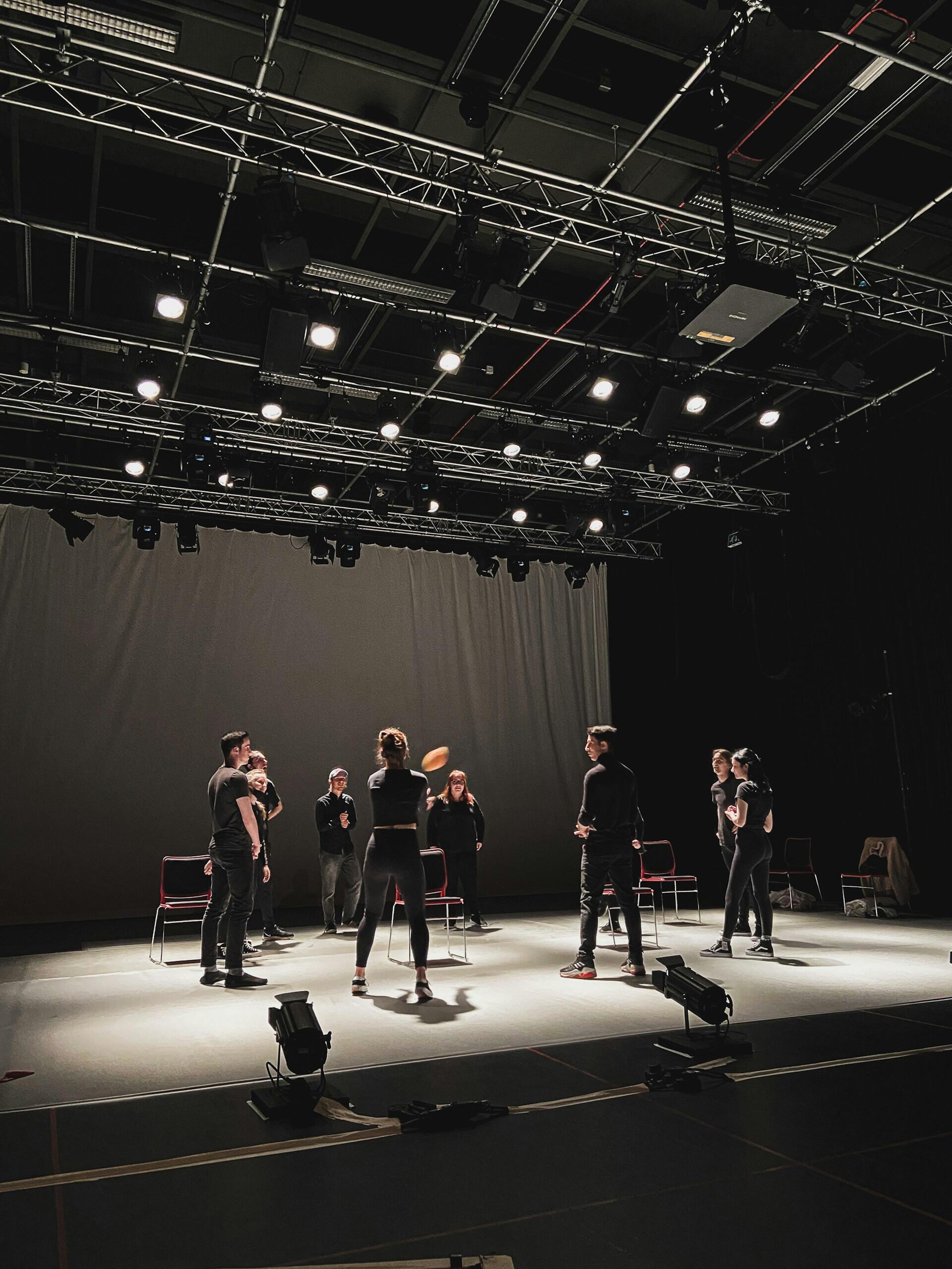 A group of performers in black attire engaging in a rehearsal on stage, surrounded by bright overhead lights and chairs.