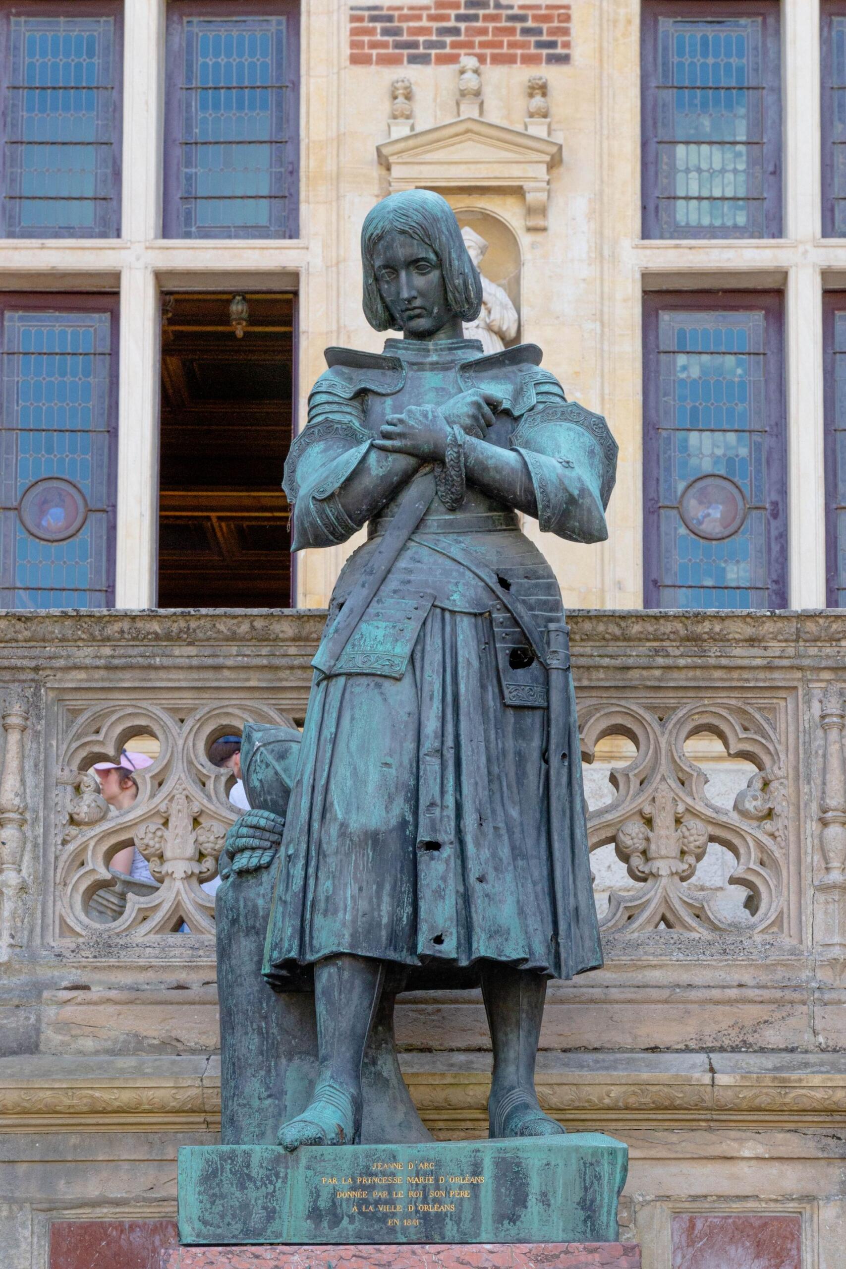 Bronze statue of Joan of Arc in armor stands in front of a building with ornate windows.