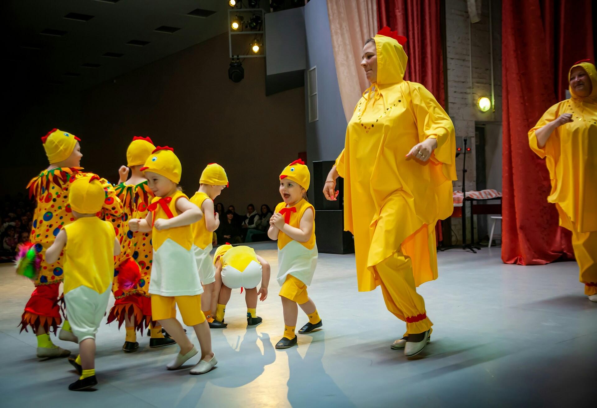 A group of children in colorful, playful costumes dance on stage, showcasing a lively performance with vibrant decorations in the background.