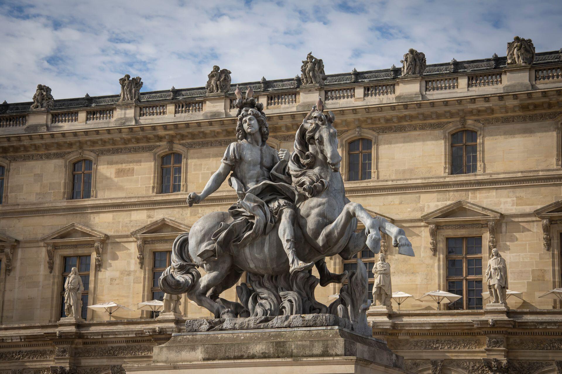 Baroque equestrian statue in front of the Louvre Museum building under a cloudy sky.