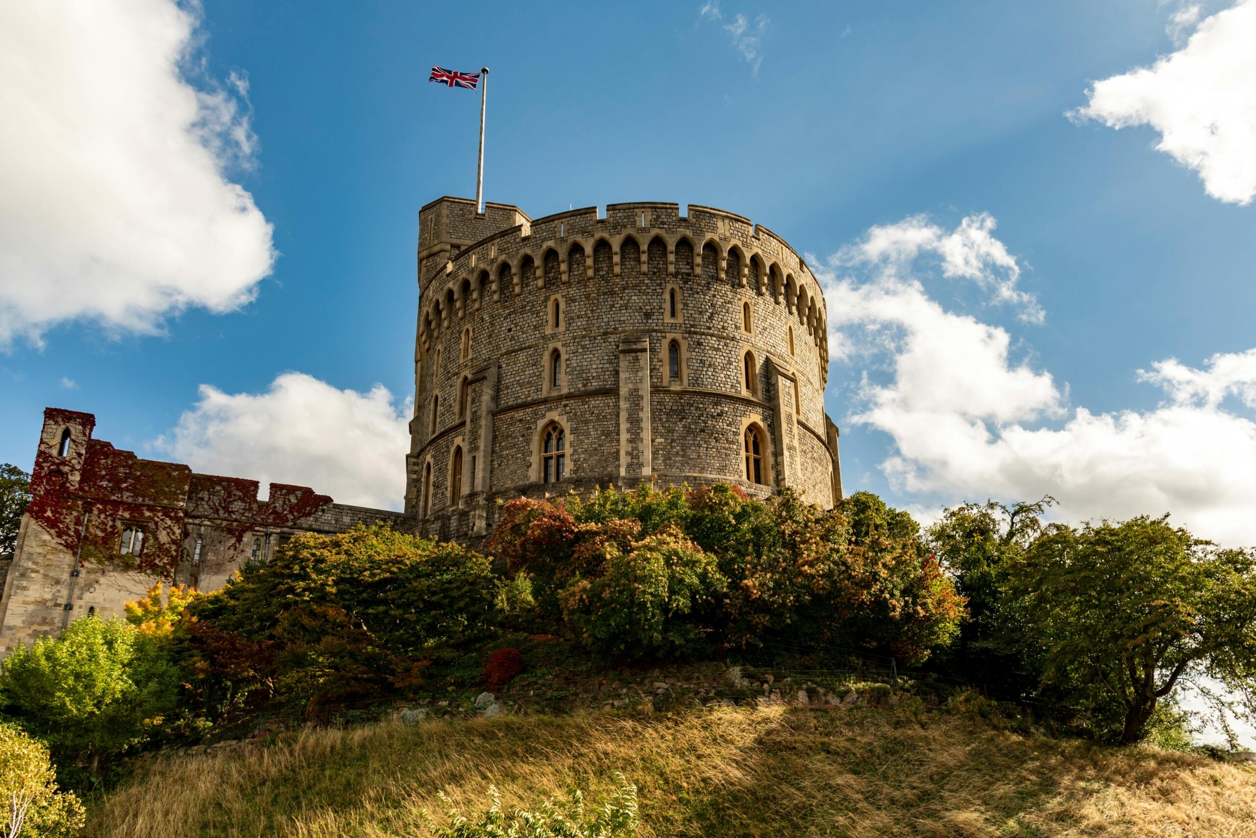 A photograph of Windsor Castle's Round Tower on a sunny day, showcasing its historic stone walls, lush green surroundings, and clear blue sky.