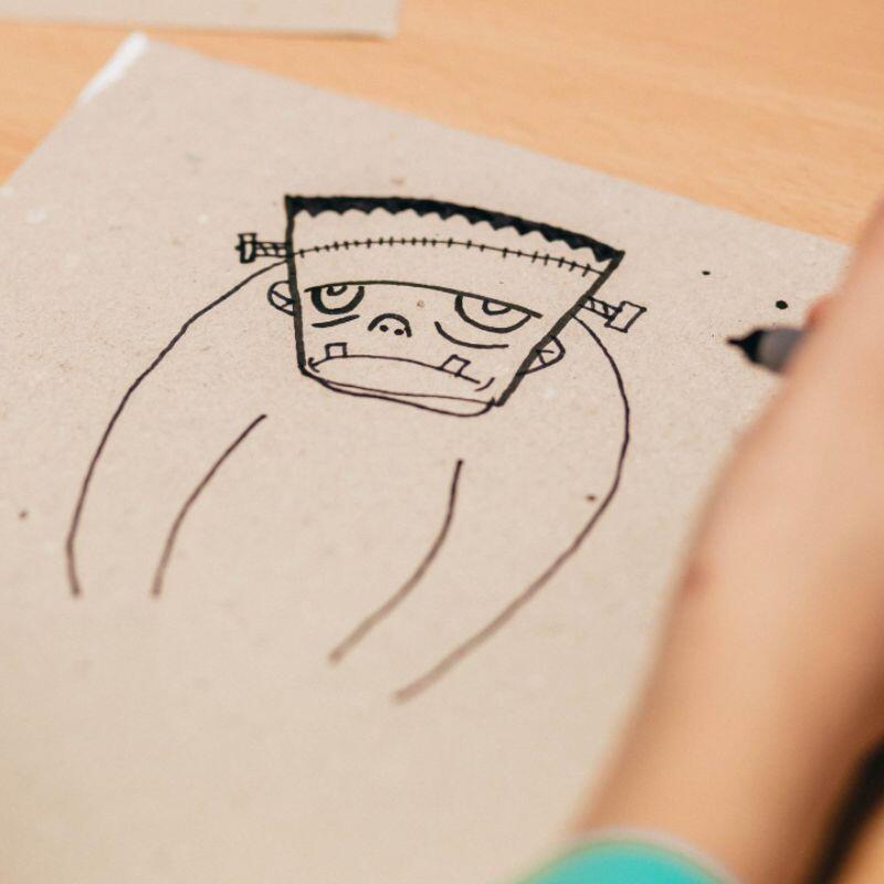 A hand drawing a cartoonish Frankenstein character on textured paper, featuring a prominent flat head and stitched facial details.