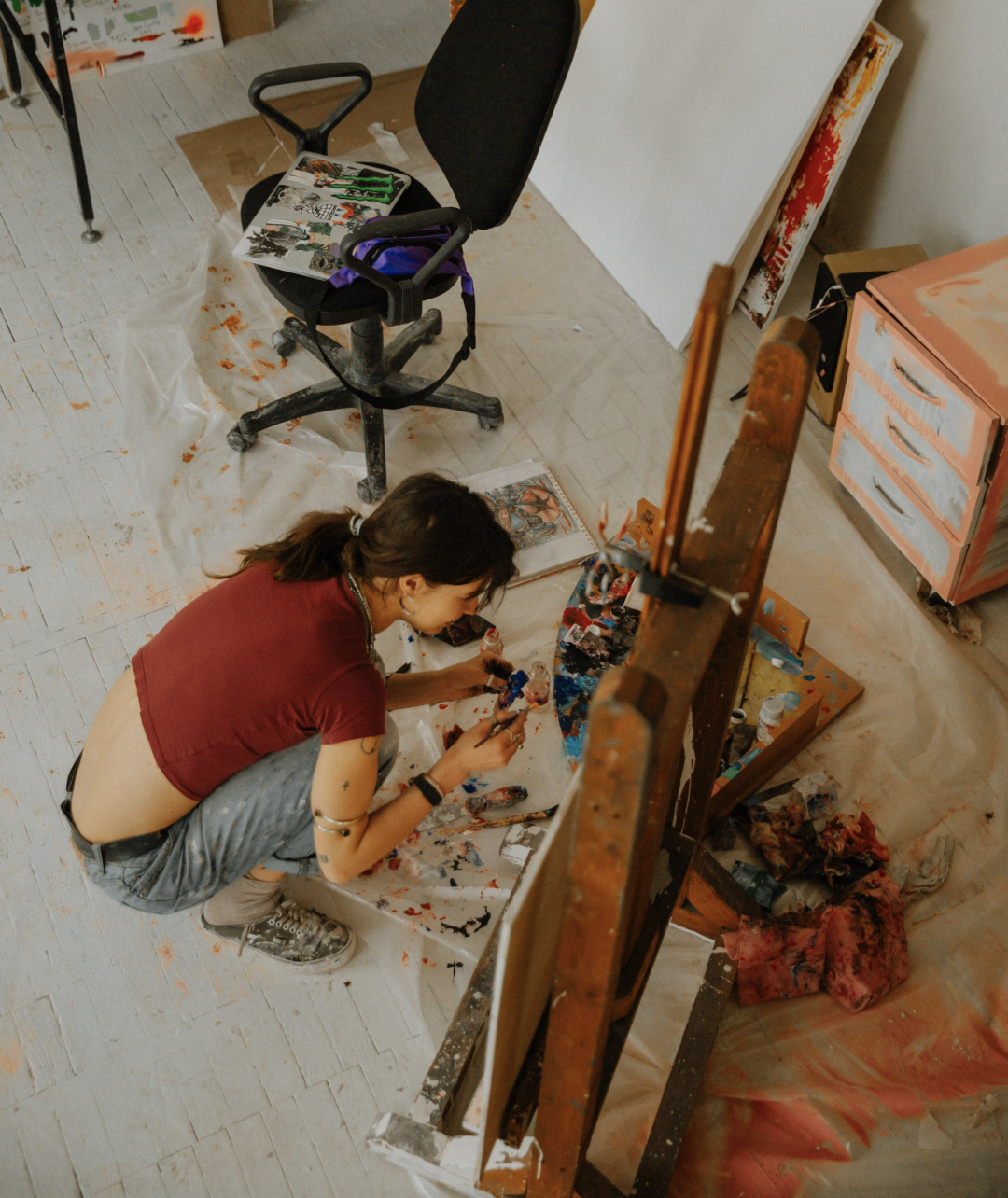 A female artist sitting in front of an easel covered in paint.