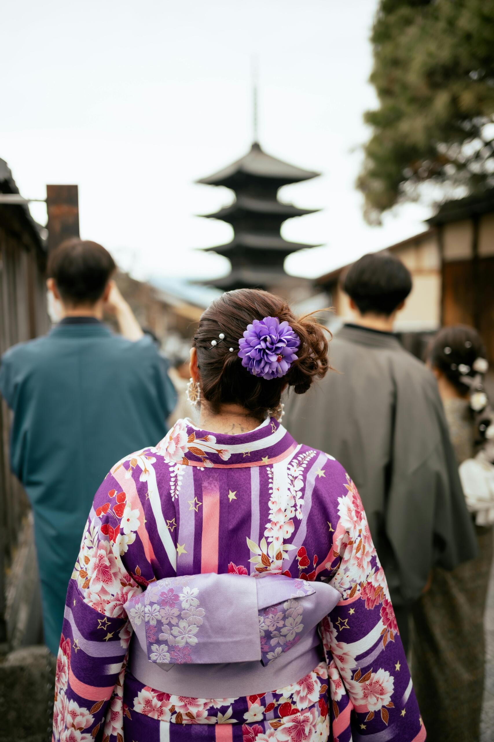 A woman dressed in traditional geisha clothing, viewed from the back, walking down a quiet street with her elaborate kimono and ornate hair accessories on full display.