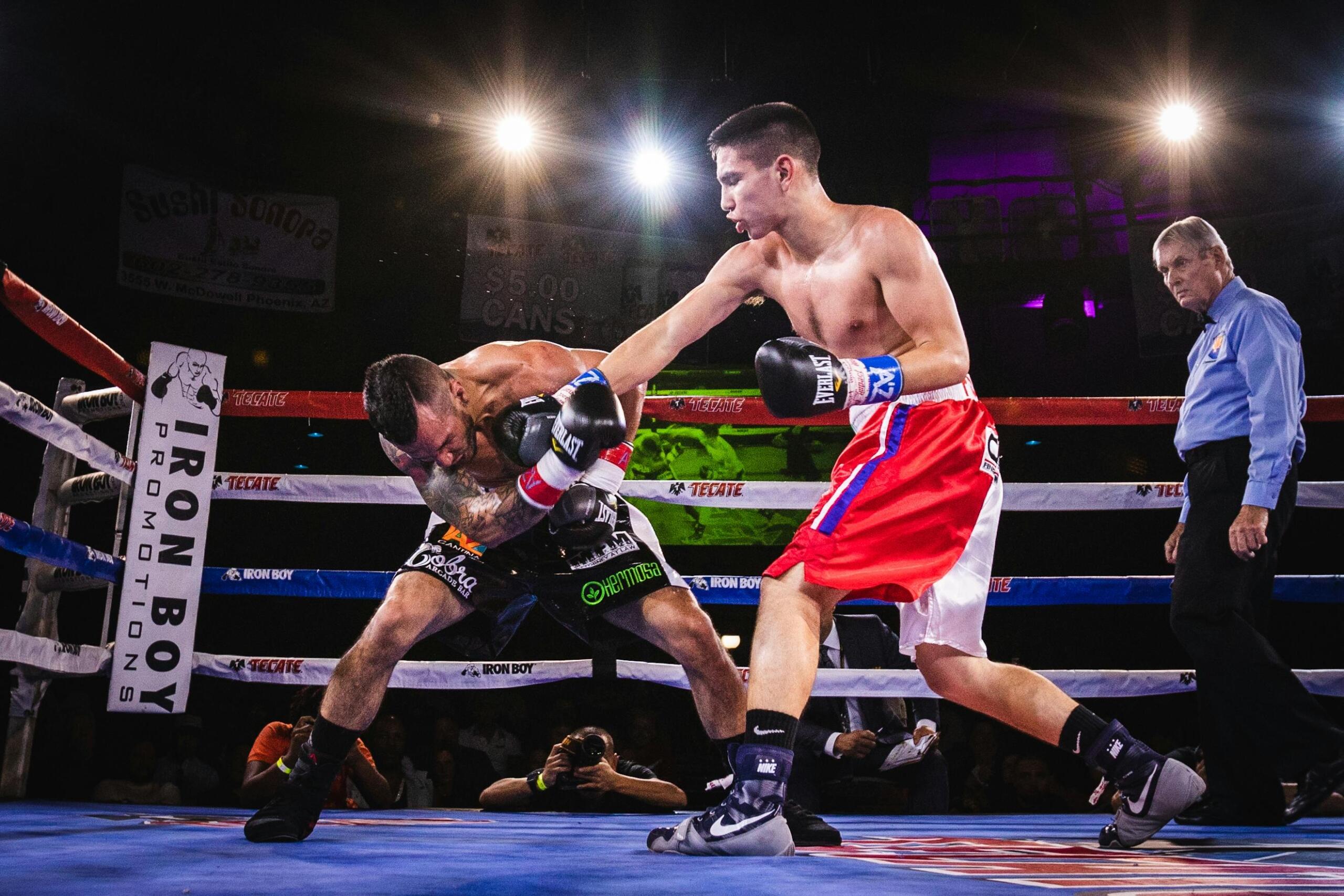 Two male boxers engage in a match inside a brightly lit boxing ring. One boxer in red and white trunks throws a punch at his opponent, who is ducking defensively near the ropes. A referee stands nearby observing the action. The scene is intense, with spectators and photographers visible in the background.