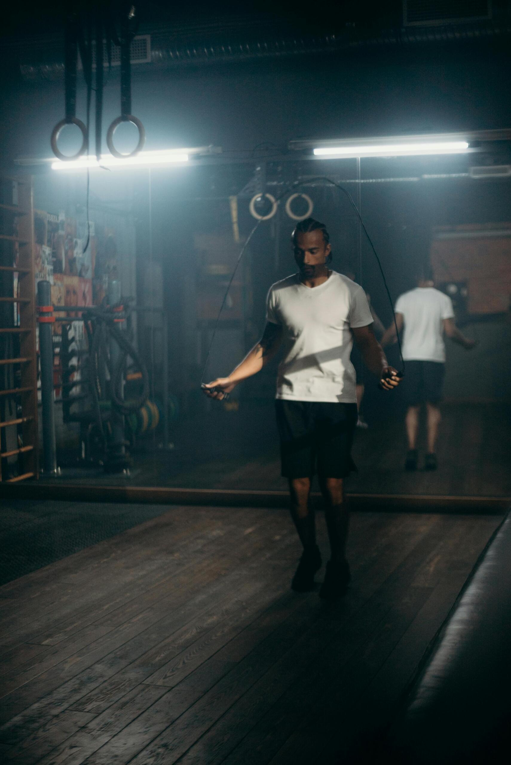 A man in a white T-shirt skips rope alone in a dimly lit gym, with his reflection visible in the mirror behind him.
