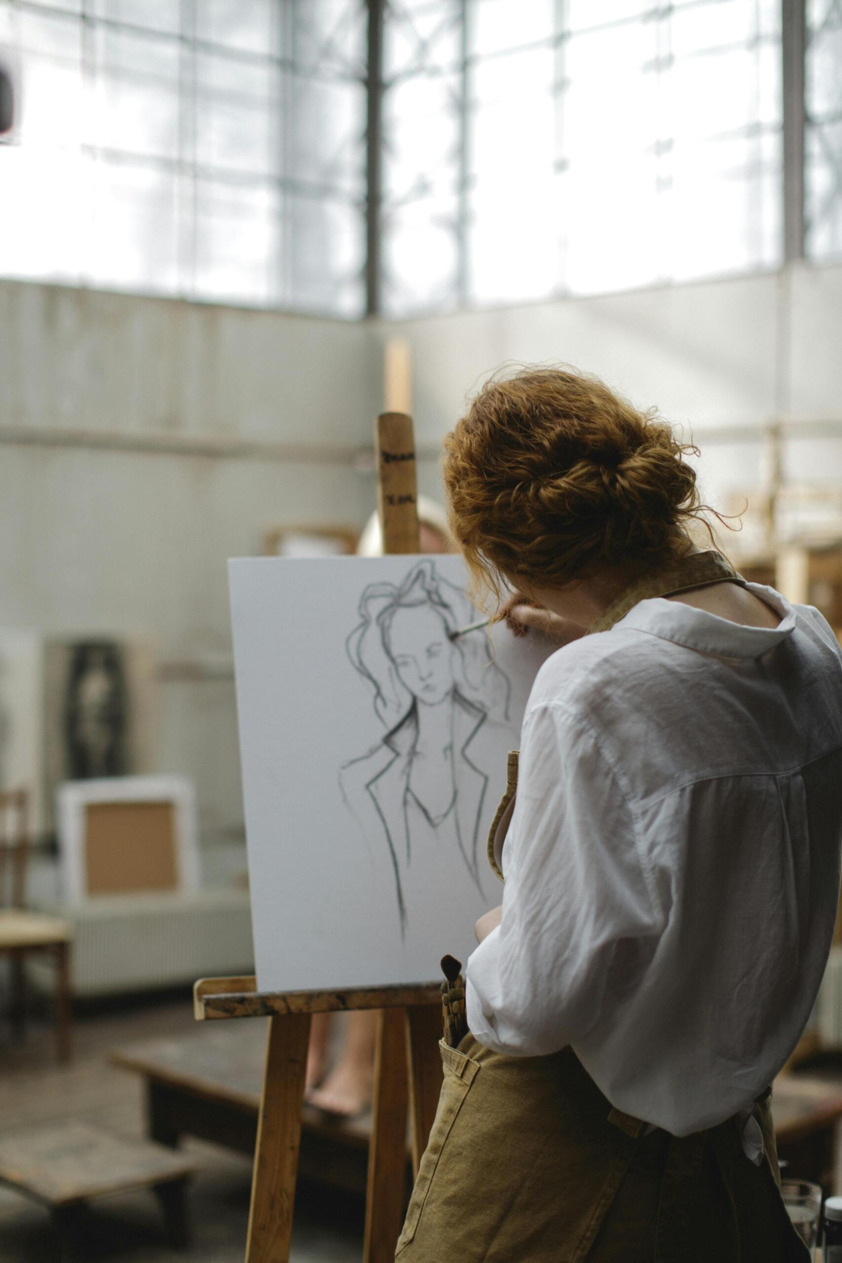 A young woman with red hair wearing an apron sketches a fashion-style portrait on a canvas in an art school studio with natural light.