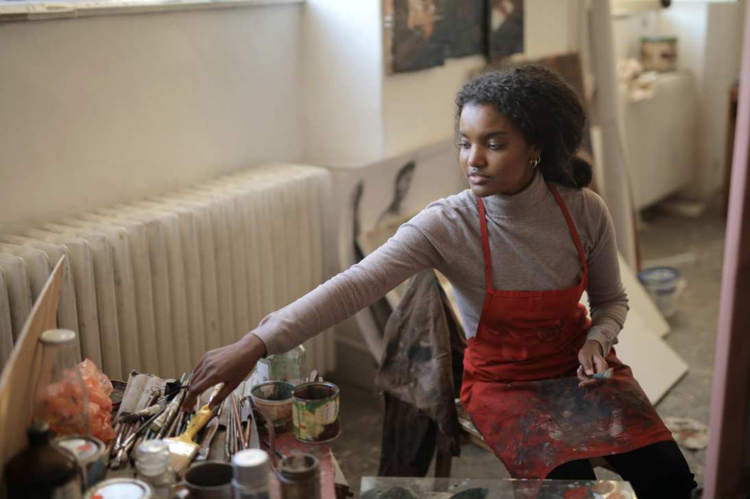 Focused art student in a red apron selects brushes in a well-used art studio.